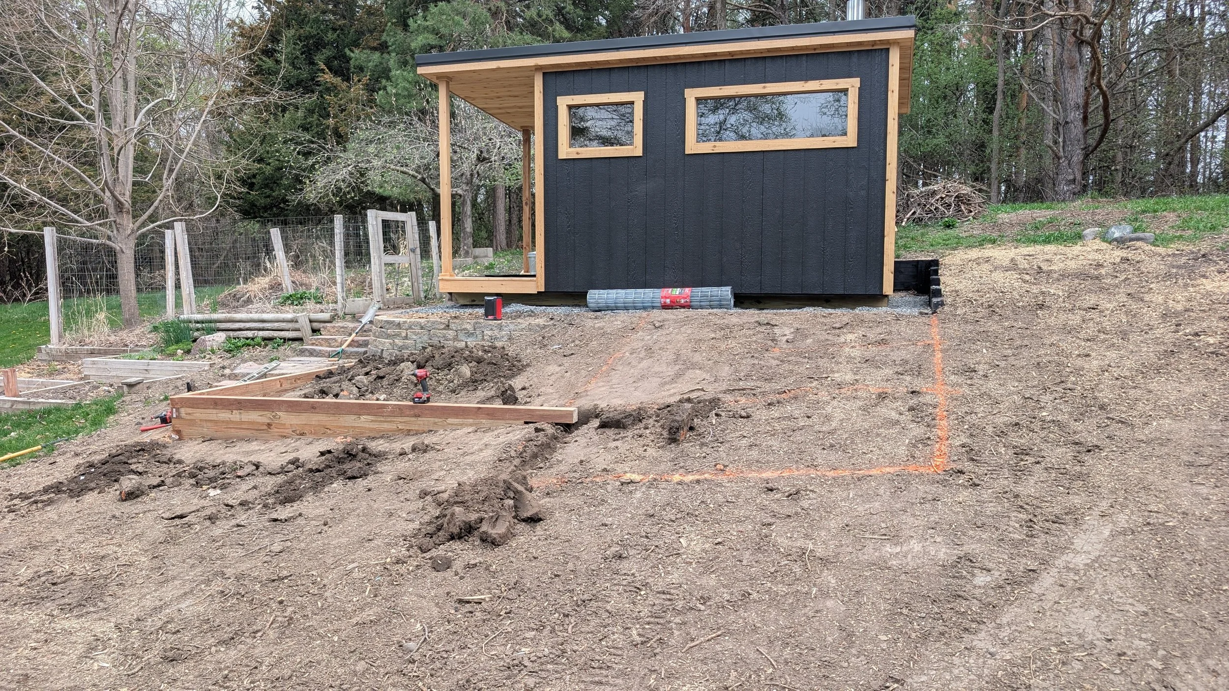 Under construction backyard with a small black shed, wooden framing, tools, and orange construction lines on dirt ground.