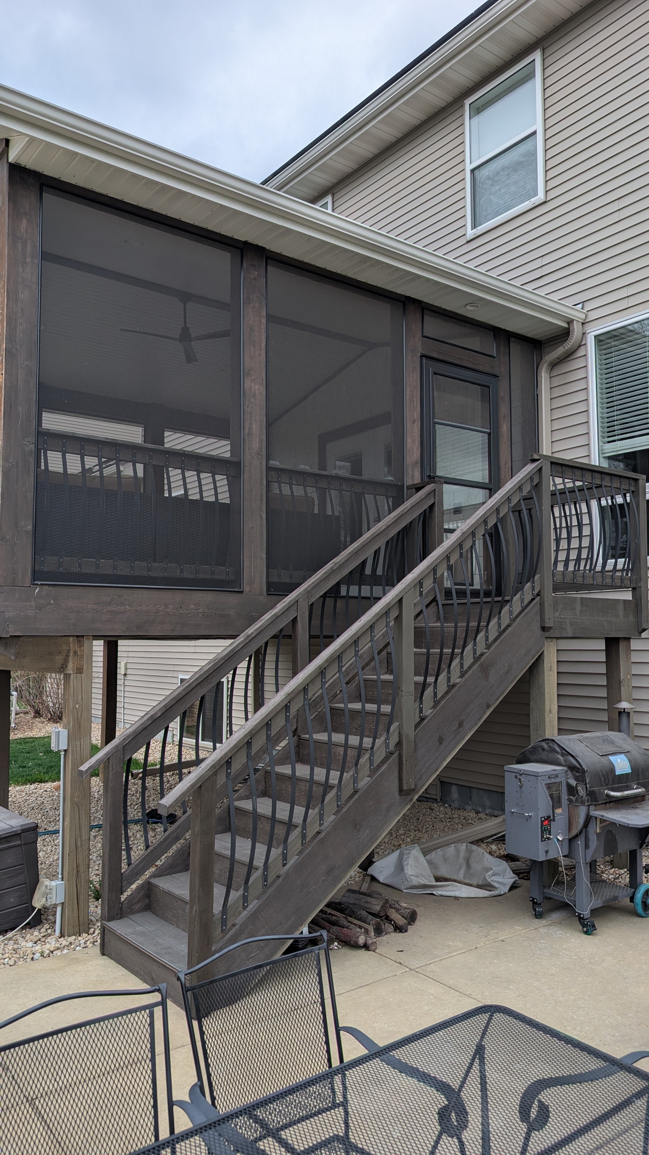 Backyard patio with a wooden staircase leading up to a screened-in porch with dark wood framing and a door. There are patio chairs and a grill on the concrete ground, with utility boxes and logs underneath.