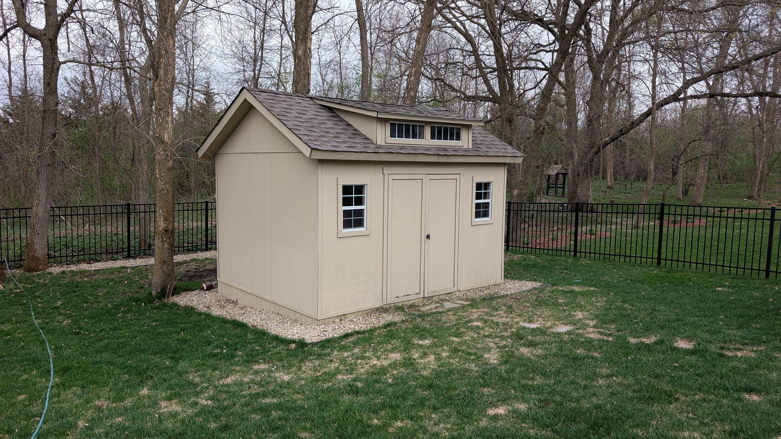 A small beige storage shed with double doors, two windows, and a shingled roof situated on a grassy yard with a black metal fence in the background, surrounded by leafless trees.