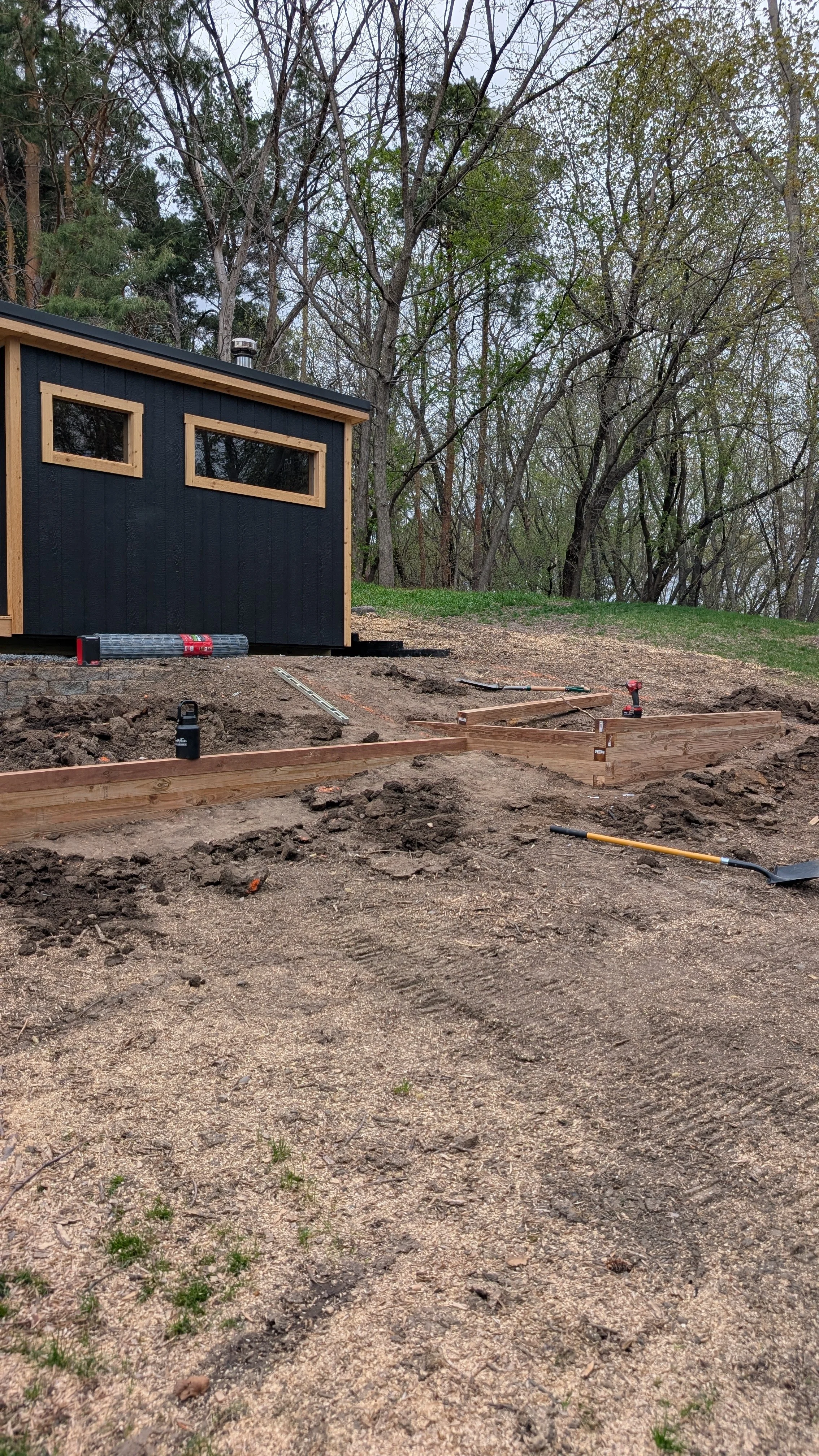Construction site with wooden framing for a small structure, dirt ground, tools, and a wooded area in the background.