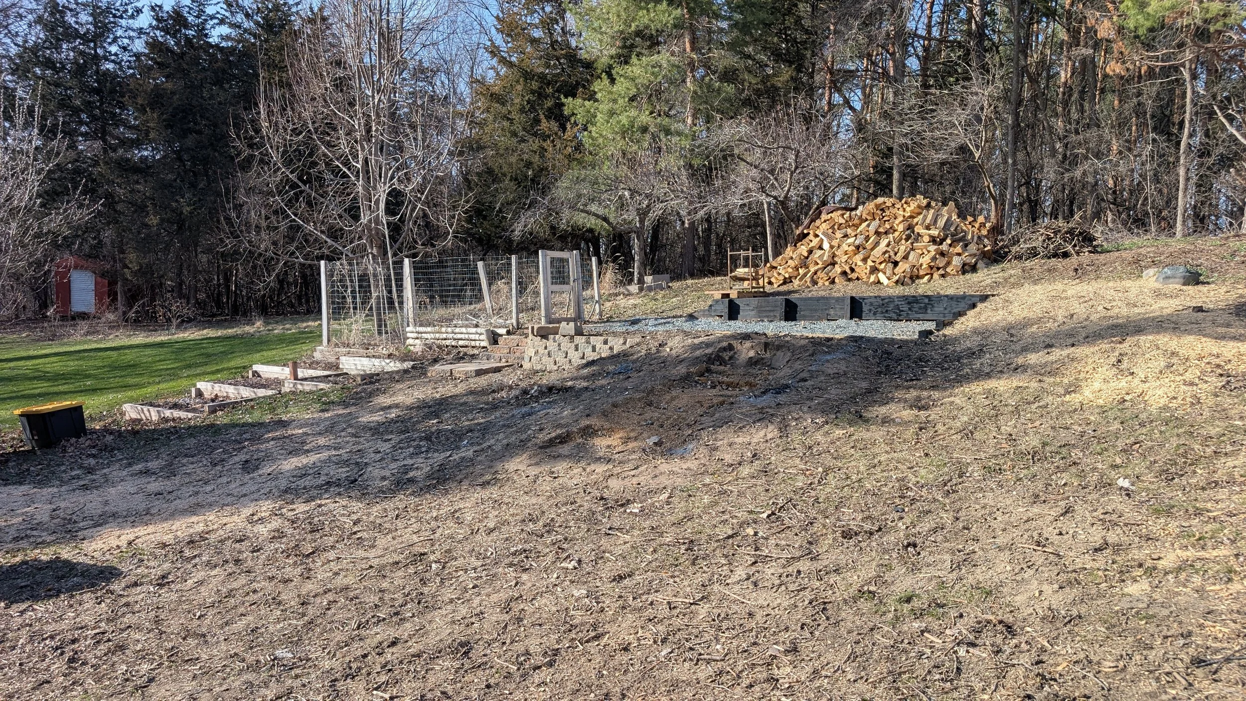 View of a backyard with a large pile of chopped firewood, a small red shed, a wood deck, a fence, and leafless trees in the background, with some evergreen trees and a clear blue sky.