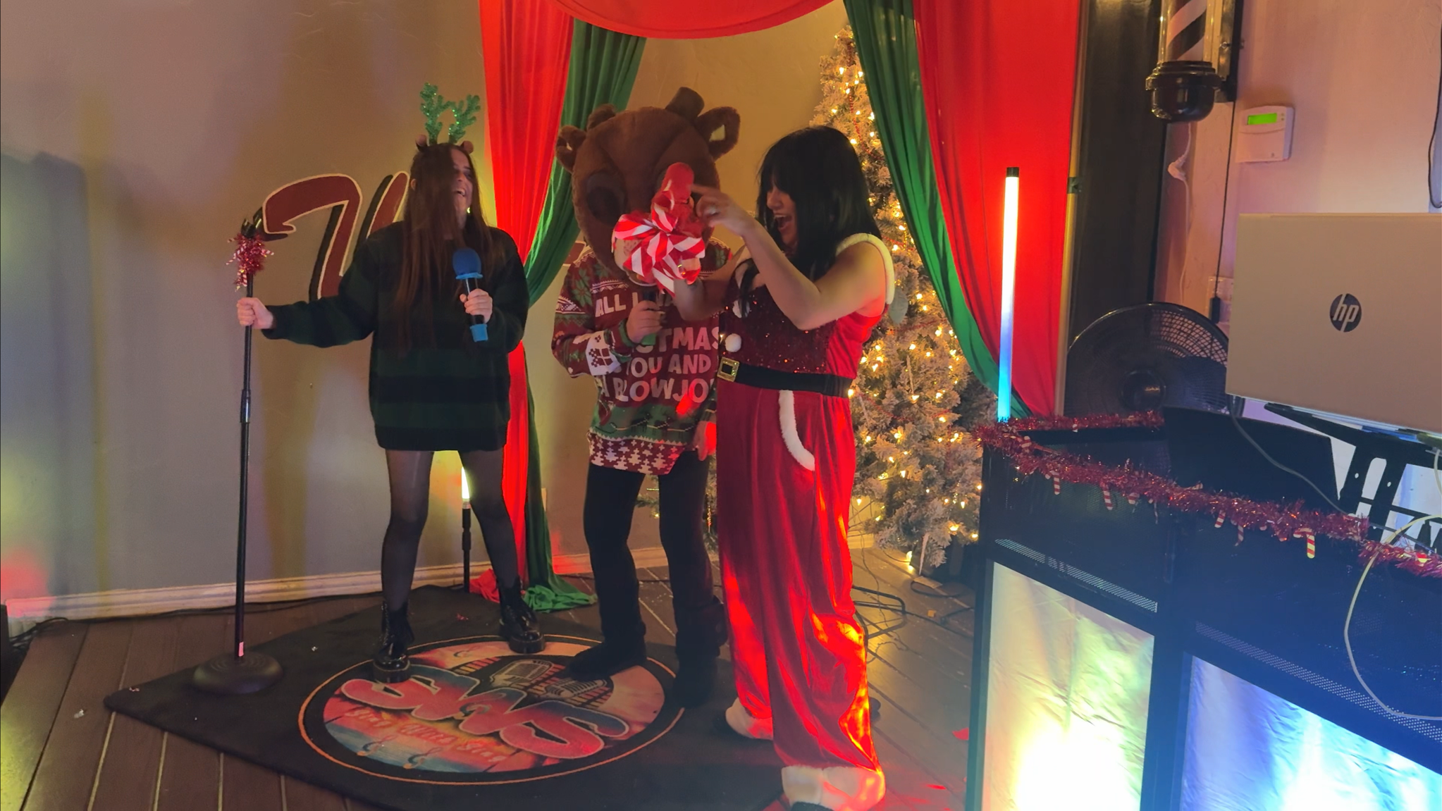 People celebrating Christmas, including a woman dressed as Santa Claus, a person in a reindeer sweater, and a woman holding a microphone, with a decorated Christmas tree and colorful curtains in the background.