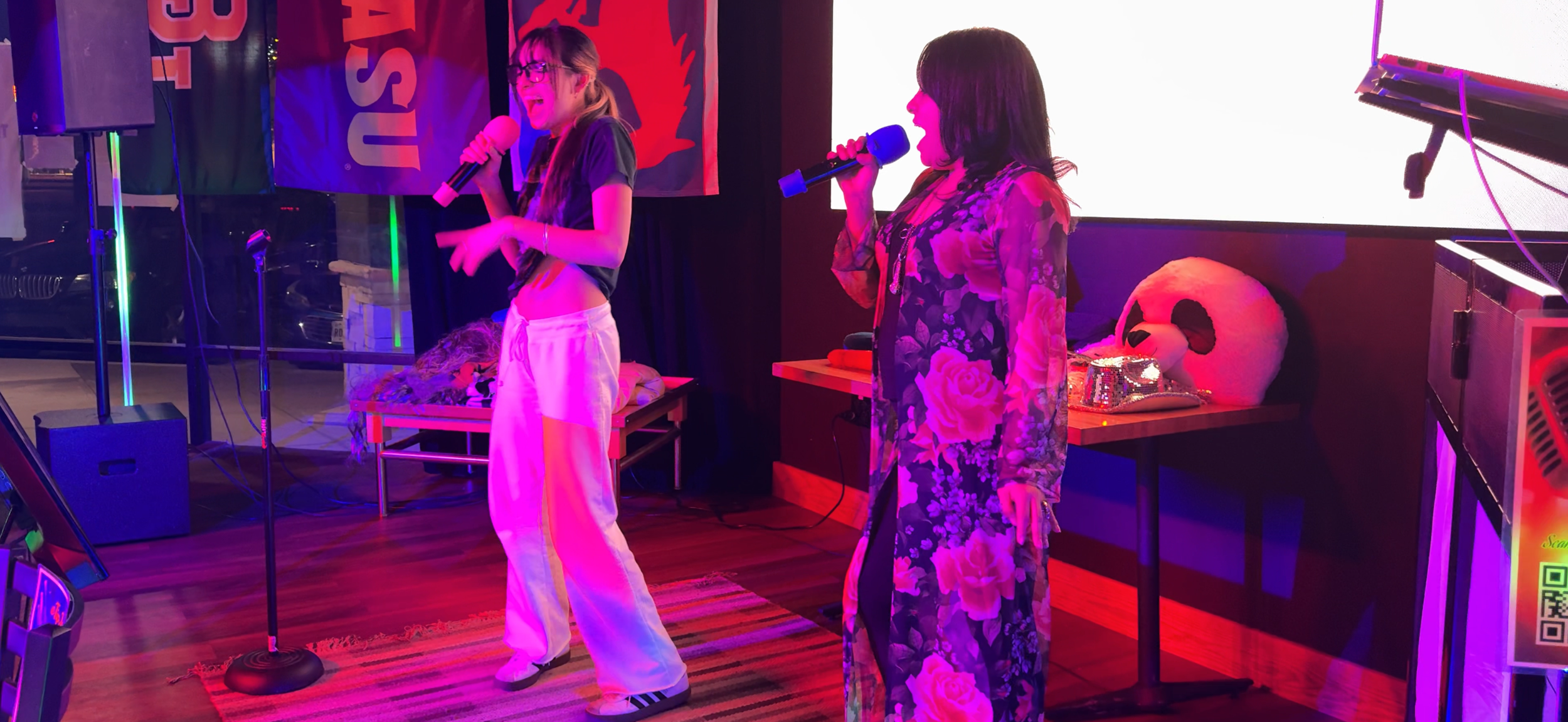 Two women singing into microphones on a small stage with neon purple lighting, colorful decorations, plush pillows, and a panda face on a pillow behind them.