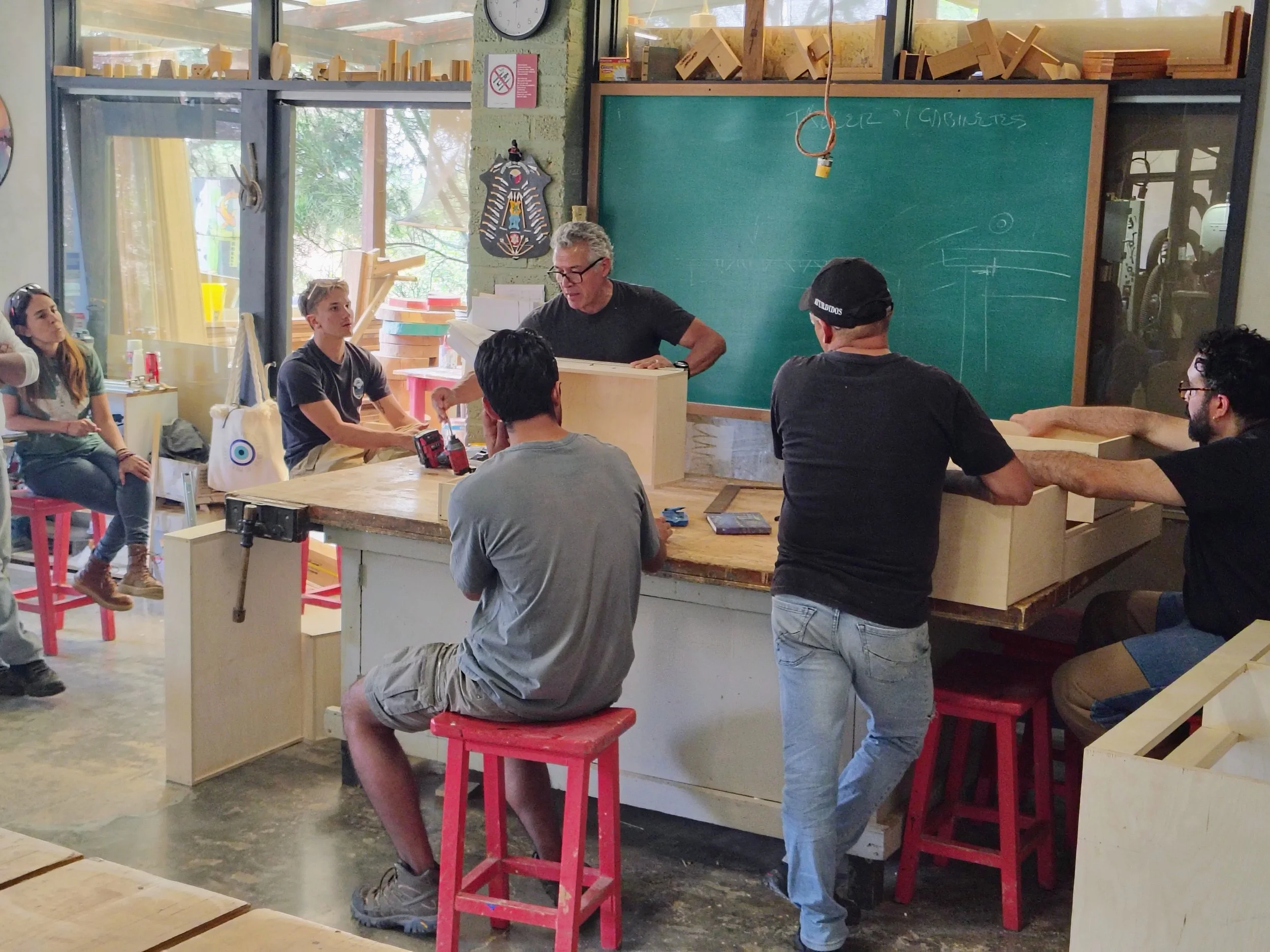 A woodworking workshop with several people, including a man with glasses at a chalkboard and others sitting on red stools, surrounded by wood pieces and tools.