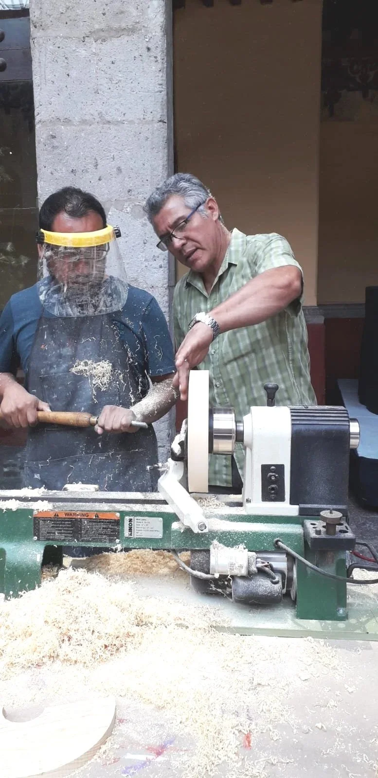 Two men working with a wood lathe in a woodworking shop. One man is wearing safety goggles and a dust mask, while the other is demonstrating or instructing.