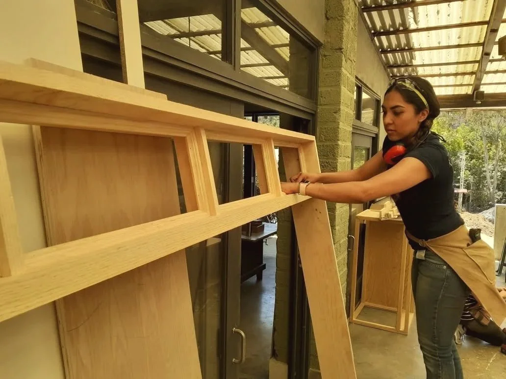 A woman working on a woodworking project inside a workshop, measuring a large wooden frame or panel.