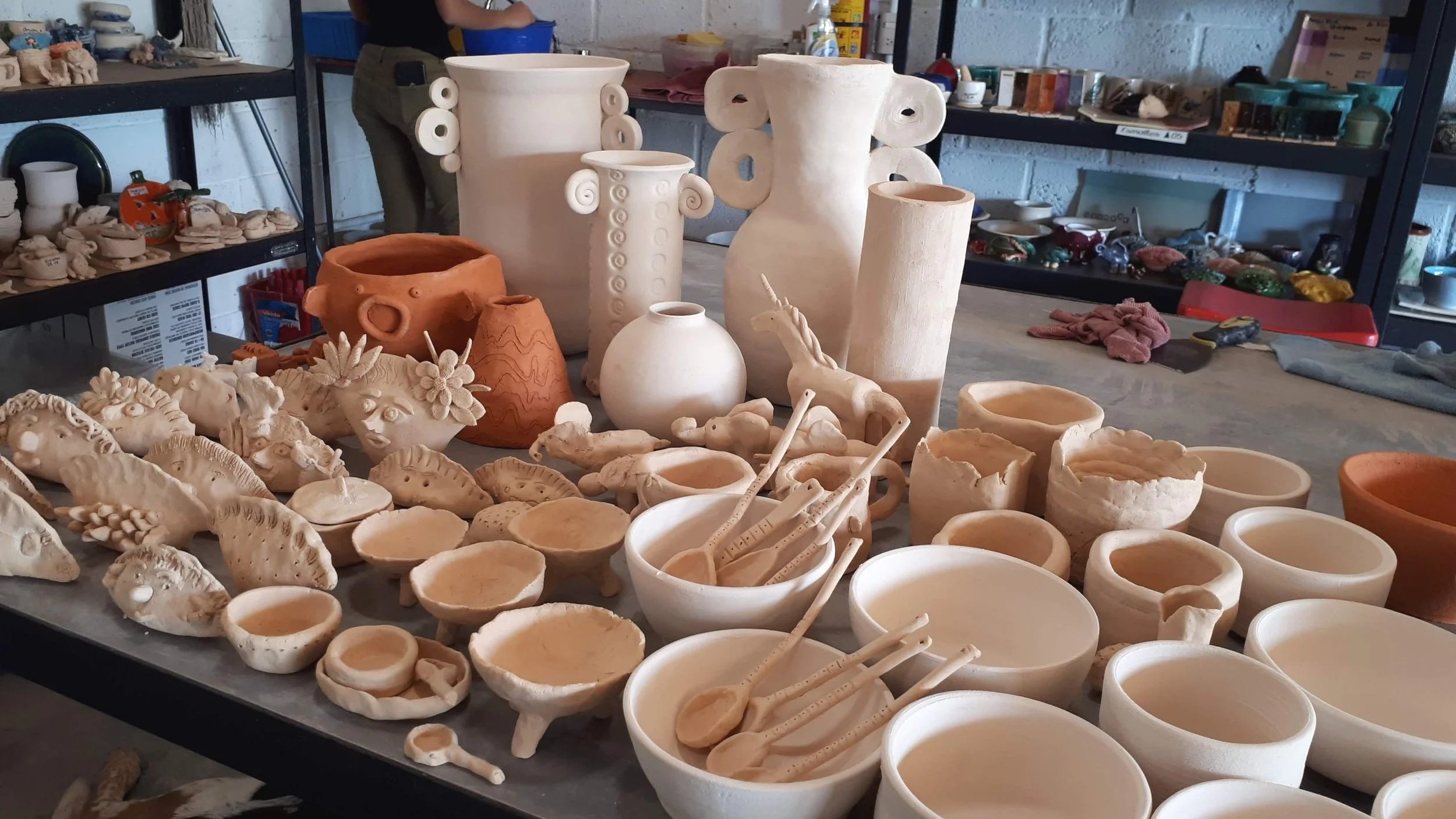 Clay sculptures and pottery pieces on a table, with shelves filled with more pottery in the background.