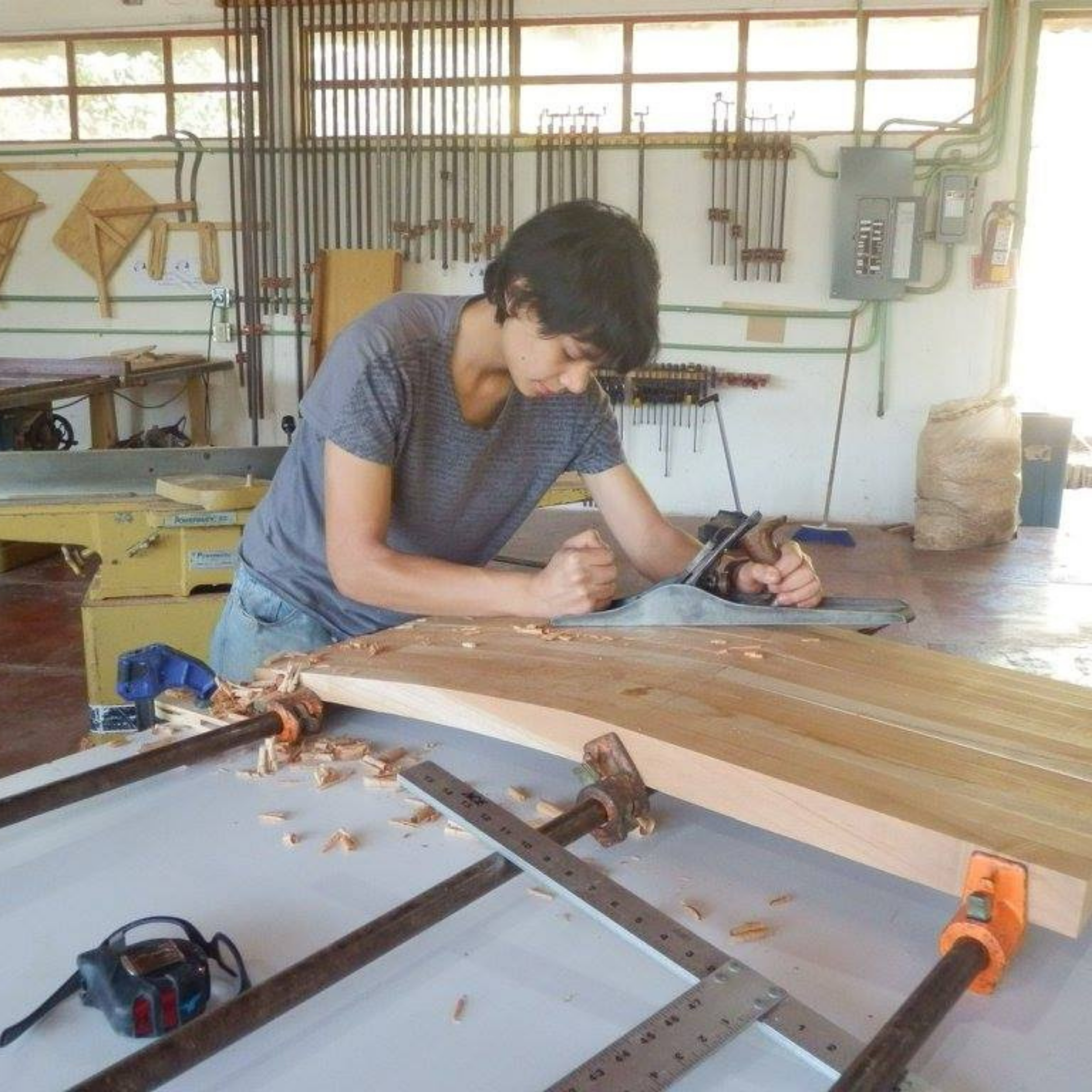 Woodworking workshop with a man cutting wood on a workbench, surrounded by tools and equipment, with a pegboard wall and electrical panel in the background.