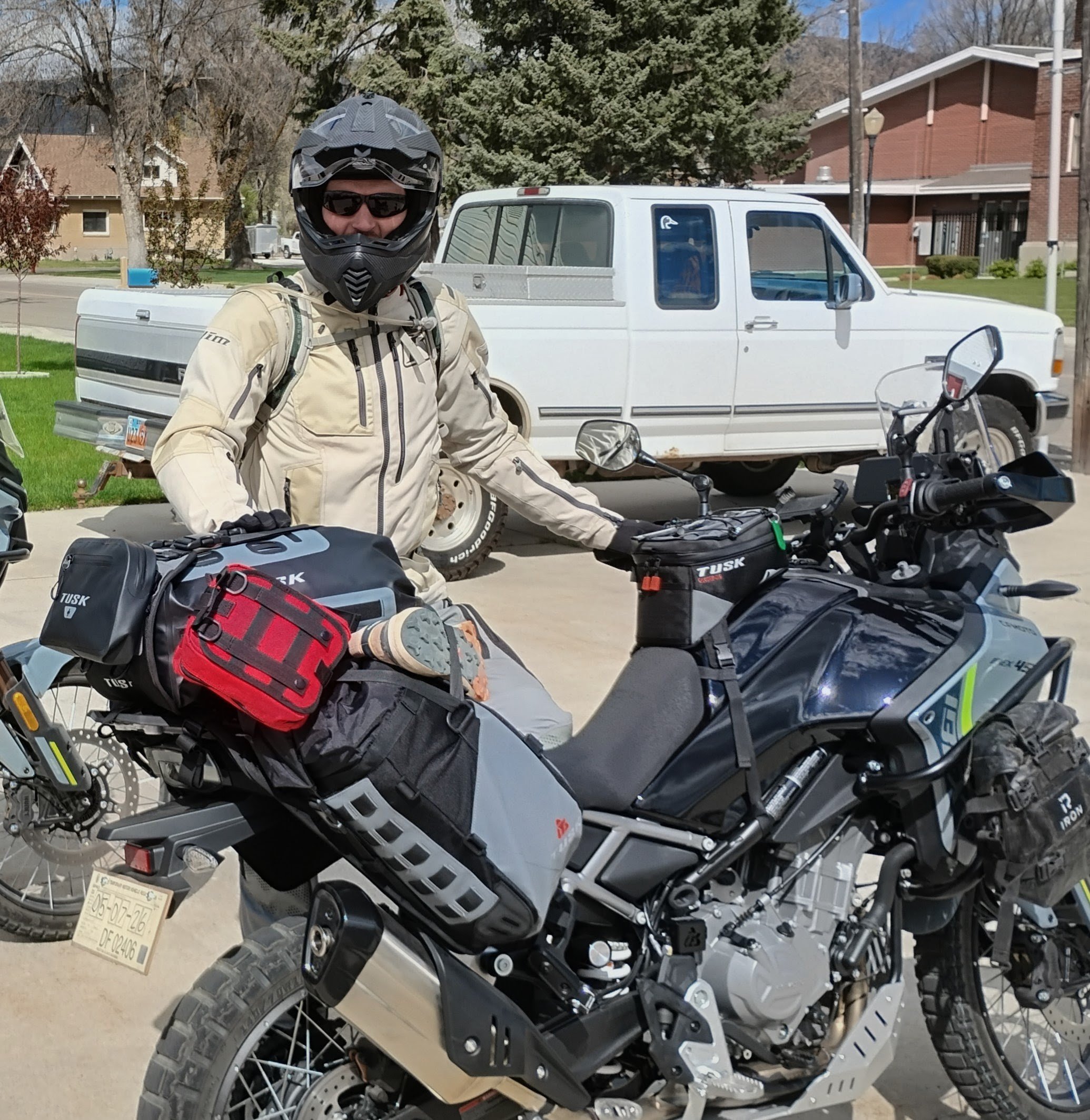 A person wearing a beige riding jacket, helmet, and sunglasses standing next to a black and white motorcycle. There is a white pickup truck and some buildings in the background under a partly cloudy sky.