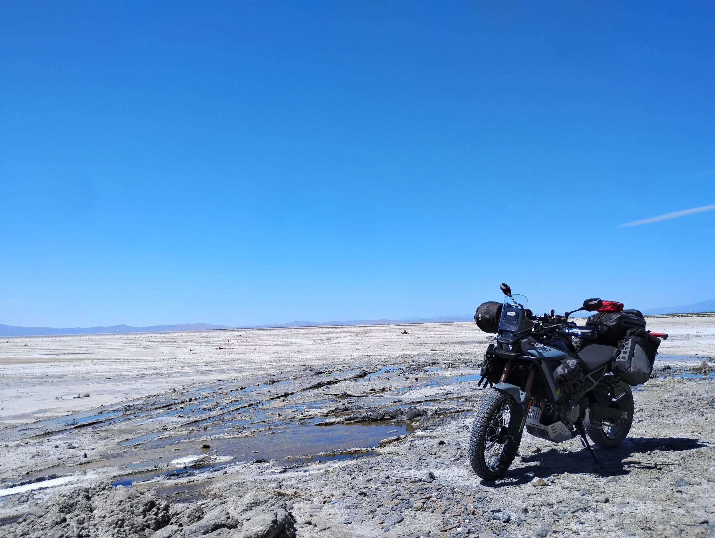 A motorcycle parked on a dry, cracked landscape with water puddles, under a clear blue sky, with mountains in the distance.