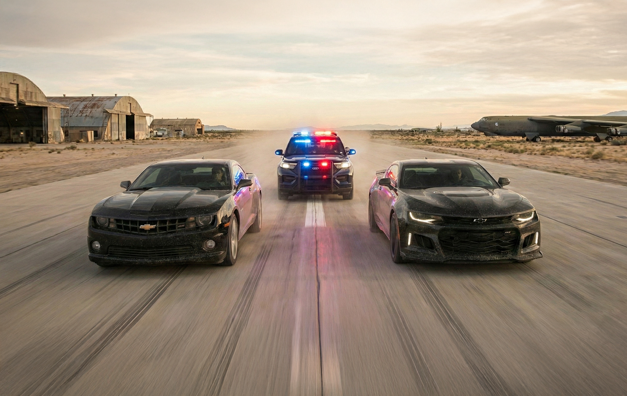 Two black police cars with flashing lights and sirens between two black sports cars on a desert runway, with scattered hangars and planes in the background.