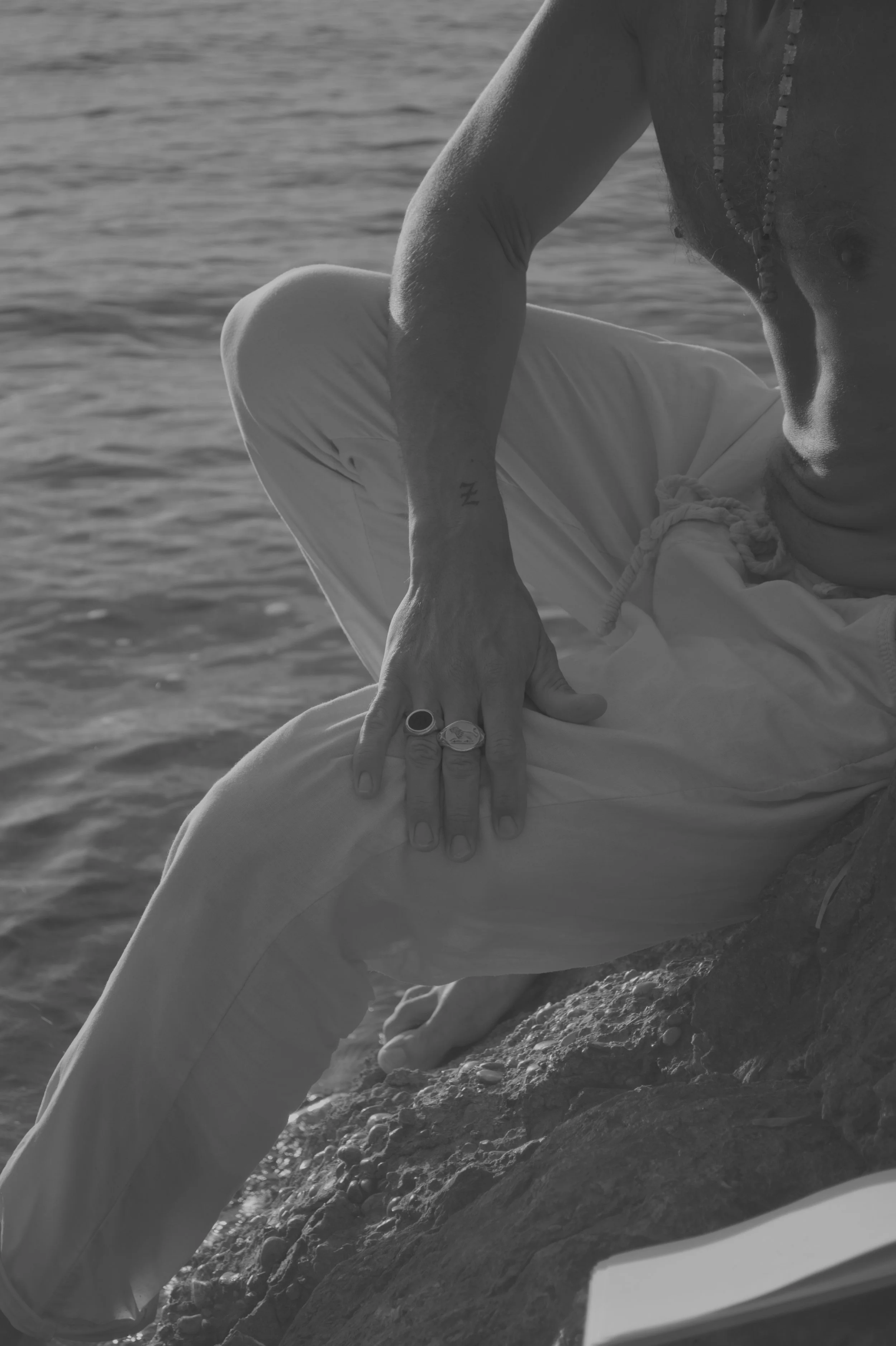 Schwarz-weiß-Foto eines Mannes, der auf einem Felsen am Wasser sitzt. Er trägt eine Halskette, eine Armbanduhr, ein Ring und eine Fußkette und scheint in einer ruhigen, natürlichen Umgebung am Wasser zu sein.