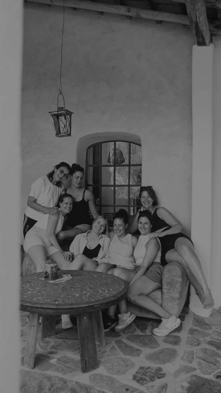 7 Frauen sitzen lächelnd auf einer urigen Couch auf einer mit Stein verzierten Außenterrasse, sie lächeln und berühren sich.
