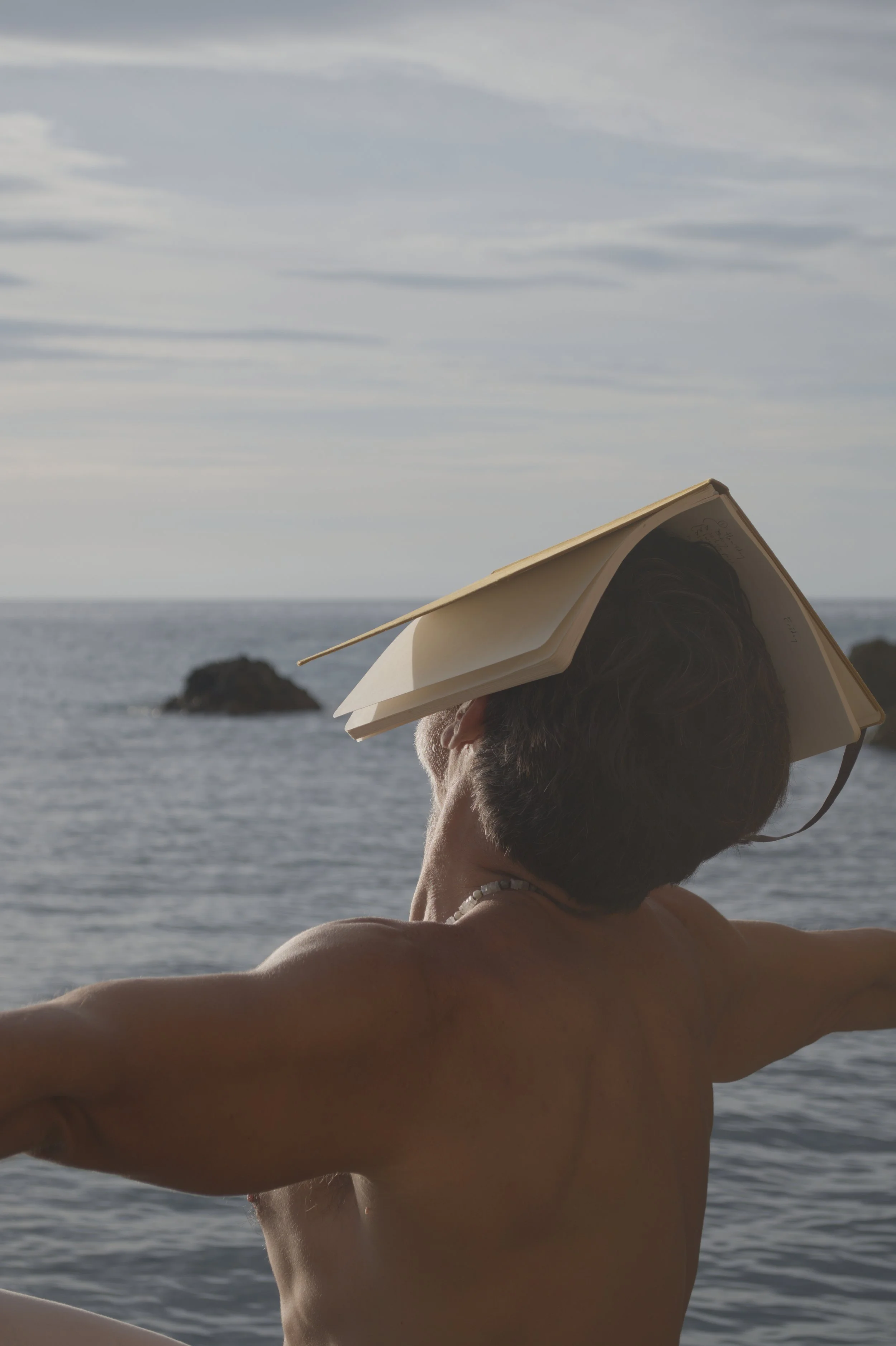 Ein Mensch mit kurzen Haaren und Muschelkette am Strand, trägt ein offenes Buch auf dem Kopf, blickt aufs Meer bei bewölktem Himmel.