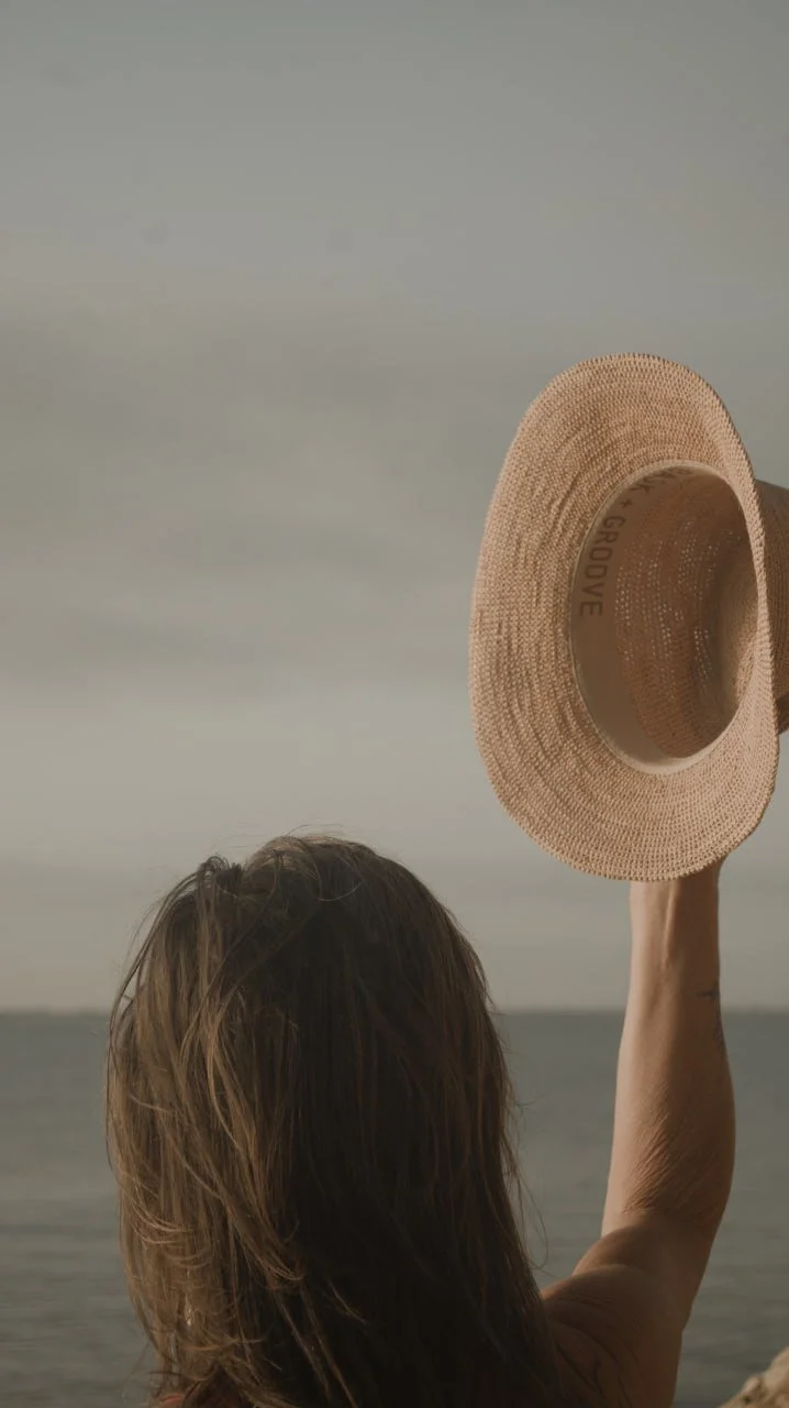 Eine Frau steht mit Blick auf das Meer an der Küste Spaniens und hält einen Hut in ihrer rechten Hand, nur ihr Hinterkopf ist zu sehen.