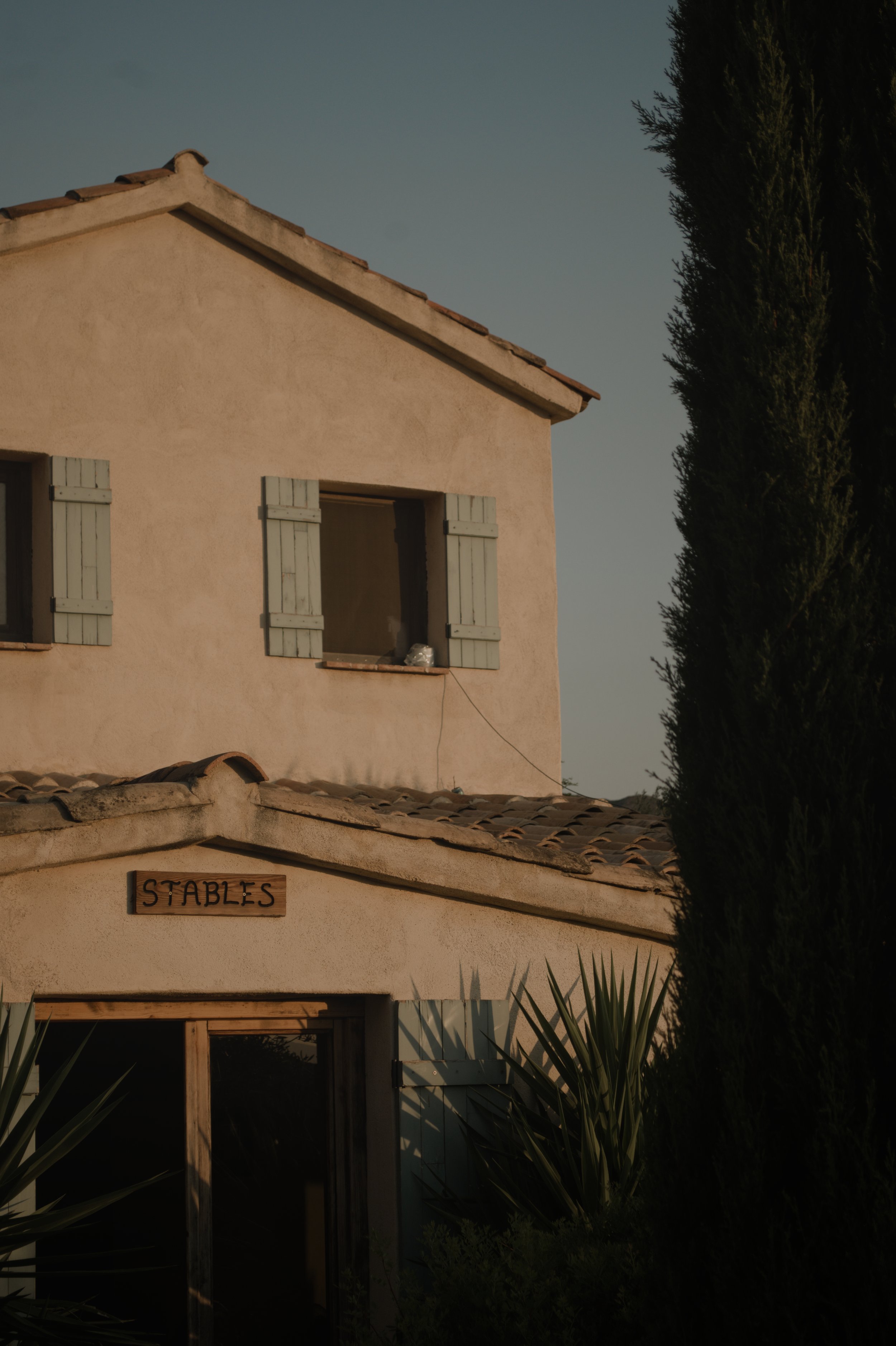 Ein zweistöckiges Haus mit hellbeigen Wänden, grünen Fensterläden und einem Schild mit der Aufschrift "Stables" an der Vorderseite, umgeben von Büschen und Bäumen.