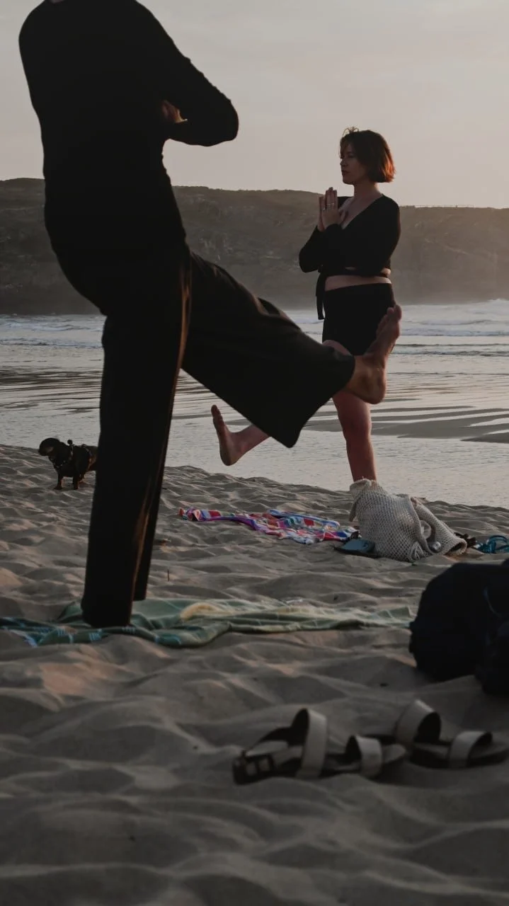 Zwei Frauen stehen sich gegenüber und praktizieren eine stehende Yogaübung im Sand, im Hintergund siehst du die Klippen Portugals