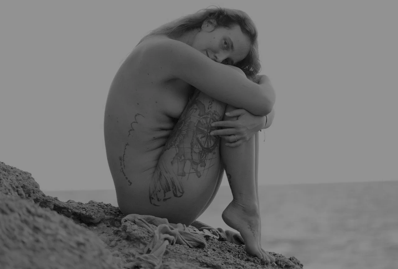Eine junge Frau sitzt nackt auf einem Felsen am Meer, ist in eine sich umarmende und liebende Haltung gehüllt und hat indianische Tattoos auf ihrem Bein und in der Rippengegend.