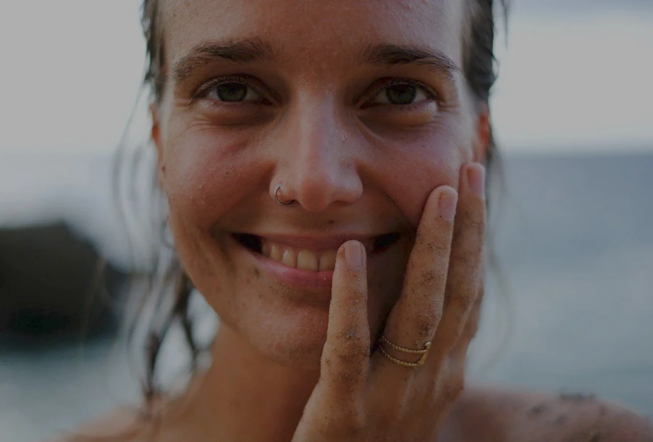 Nahaufnahme einer lächelnden Frau mit nassem Haar, Wasser im Hintergrund, trägt einen Nasenring und mehrere Goldringe an den Fingern, die sich an ihrem Gesicht berühren.
