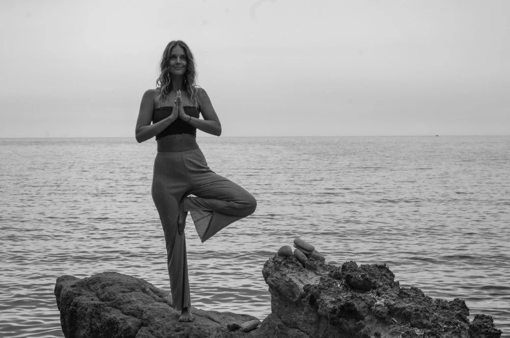 Frau, die auf einem Felsen am Meer in einem Baum-Haltung steht, die Hände vor der Brust zusammengefügt.