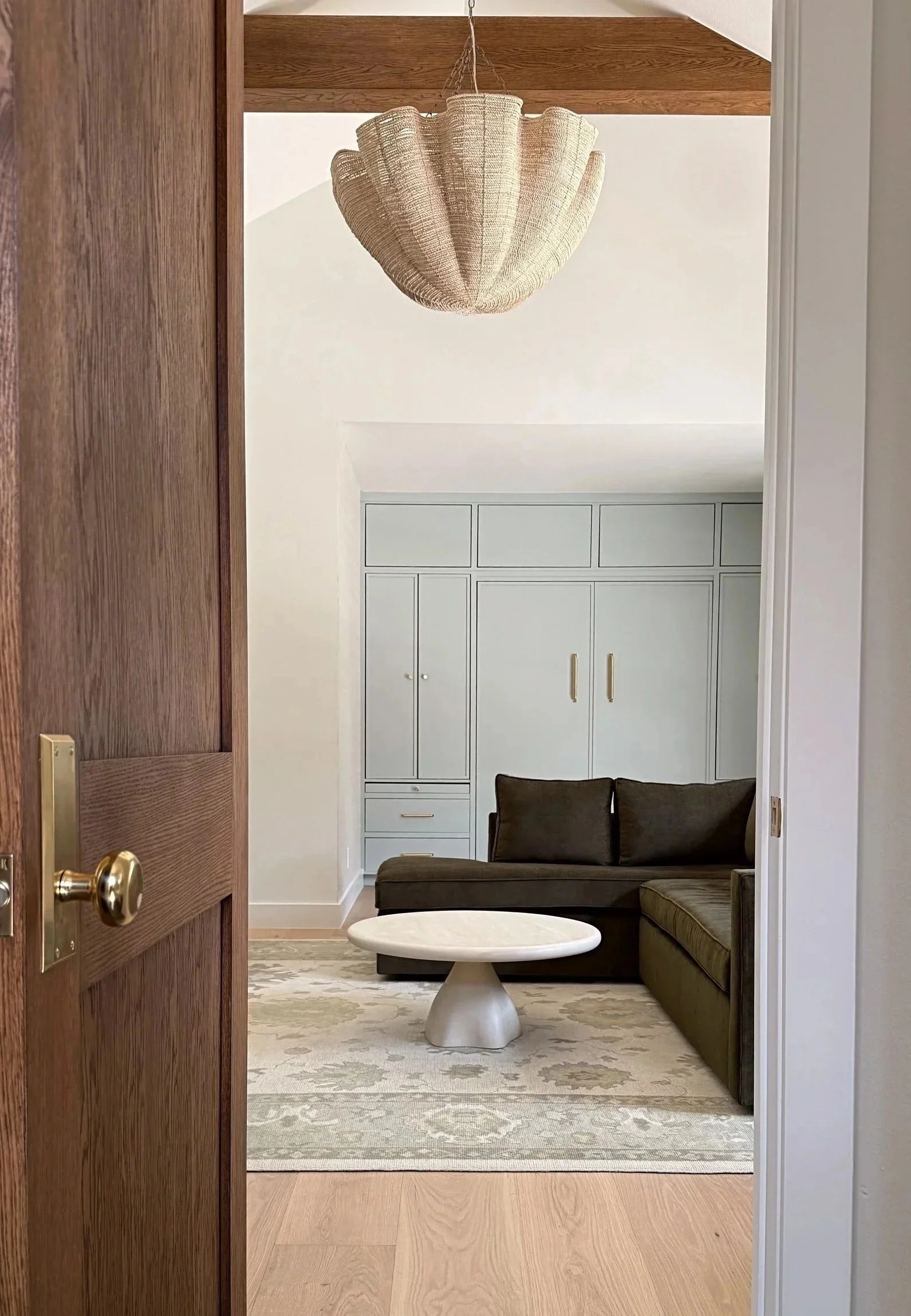 A doorway opens into a cozy living room with a dark green sectional sofa, a small white marble-topped table, and built-in cabinets painted in a muted light green color. The room features a patterned area rug, and a pendant light with a woven, beige lampshade hangs from the ceiling. There is wood paneling on the ceiling near the entrance.