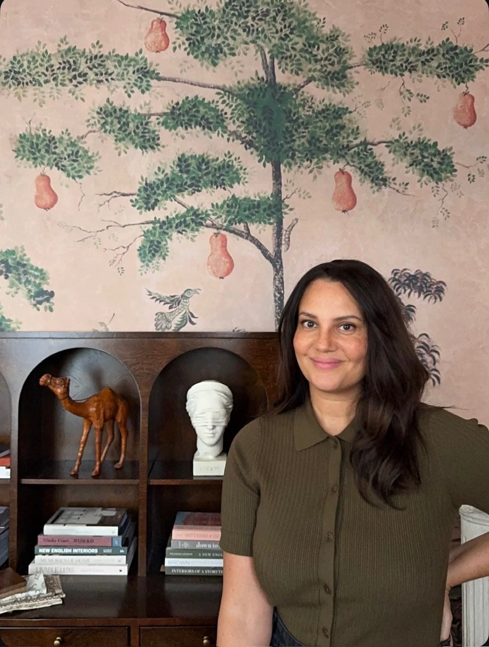 A woman with long dark hair smiling in front of a wall mural featuring a tree with hanging pears, birds,  with shelves holding books, a camel sculpture, and a beautiful female sculpture.