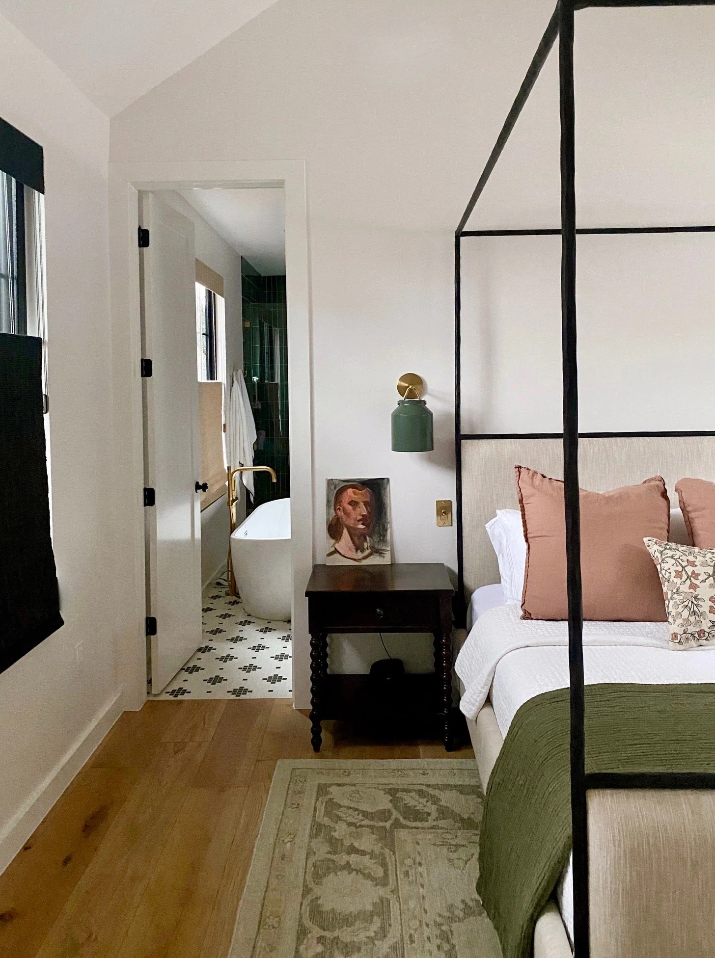 A bedroom with a cream-colored bed, featuring pink and patterned pillows, a dark wooden nightstand, a green bedspread, and a black metal canopy frame. A white curtain with floral accents hangs on the bed. To the left, a white door leads to an en suite bathroom with green tiles and a bathtub, visible through the doorway. The bathroom has two windows, one with a beige shade. There's a painting of a woman with red hair on the wall above the nightstand, and a green wall-mounted lamp is attached above the painting. The room has light hardwood floors, a beige and gray area rug, and a black-and-white tiled floor in the bathroom.