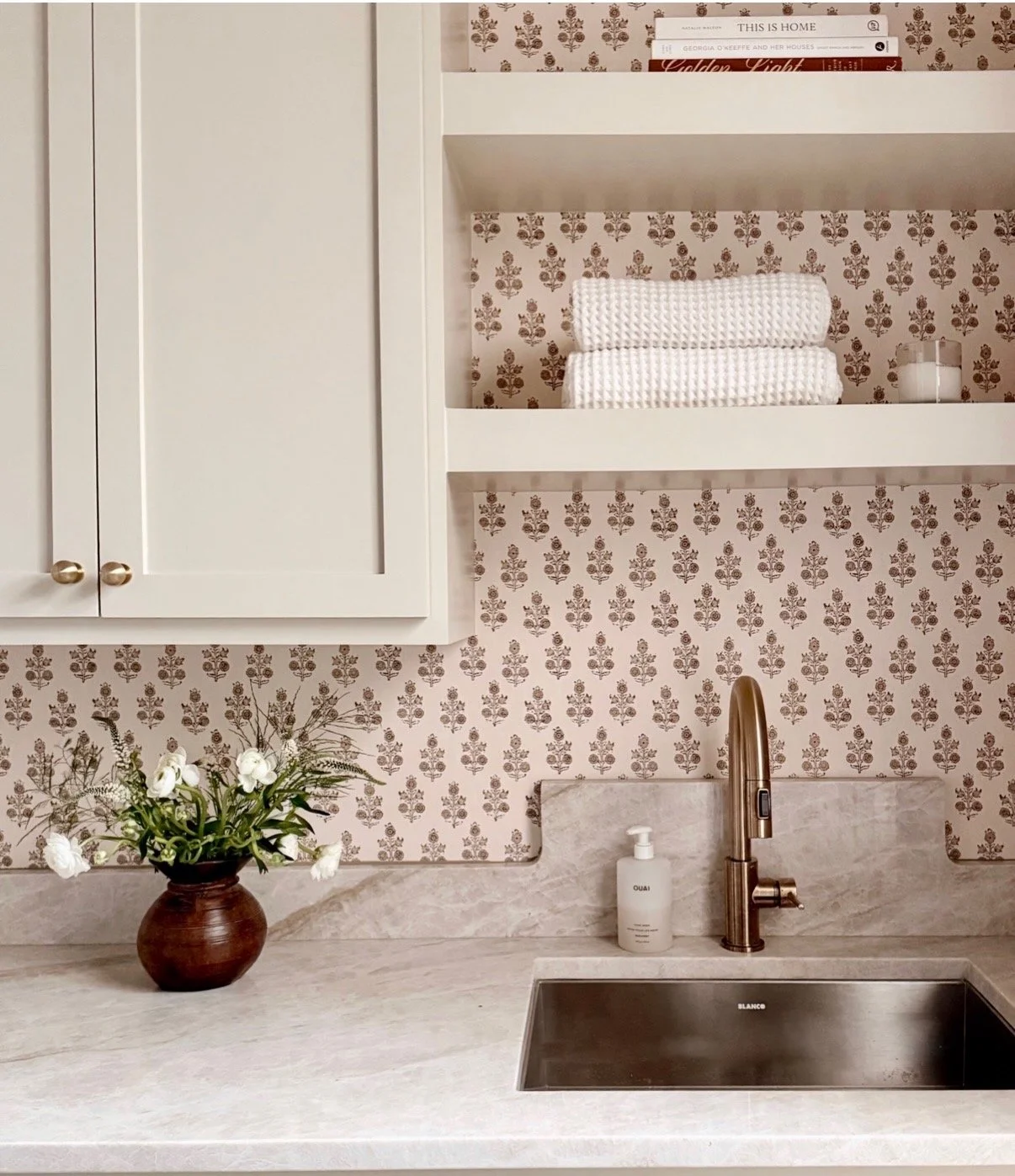 A kitchen countertop with a flower vase, soap dispenser, and a sink with a bronze faucet. The backsplash has a small floral pattern wallpaper, and above are white open shelves with books, folded towels, and a candle.