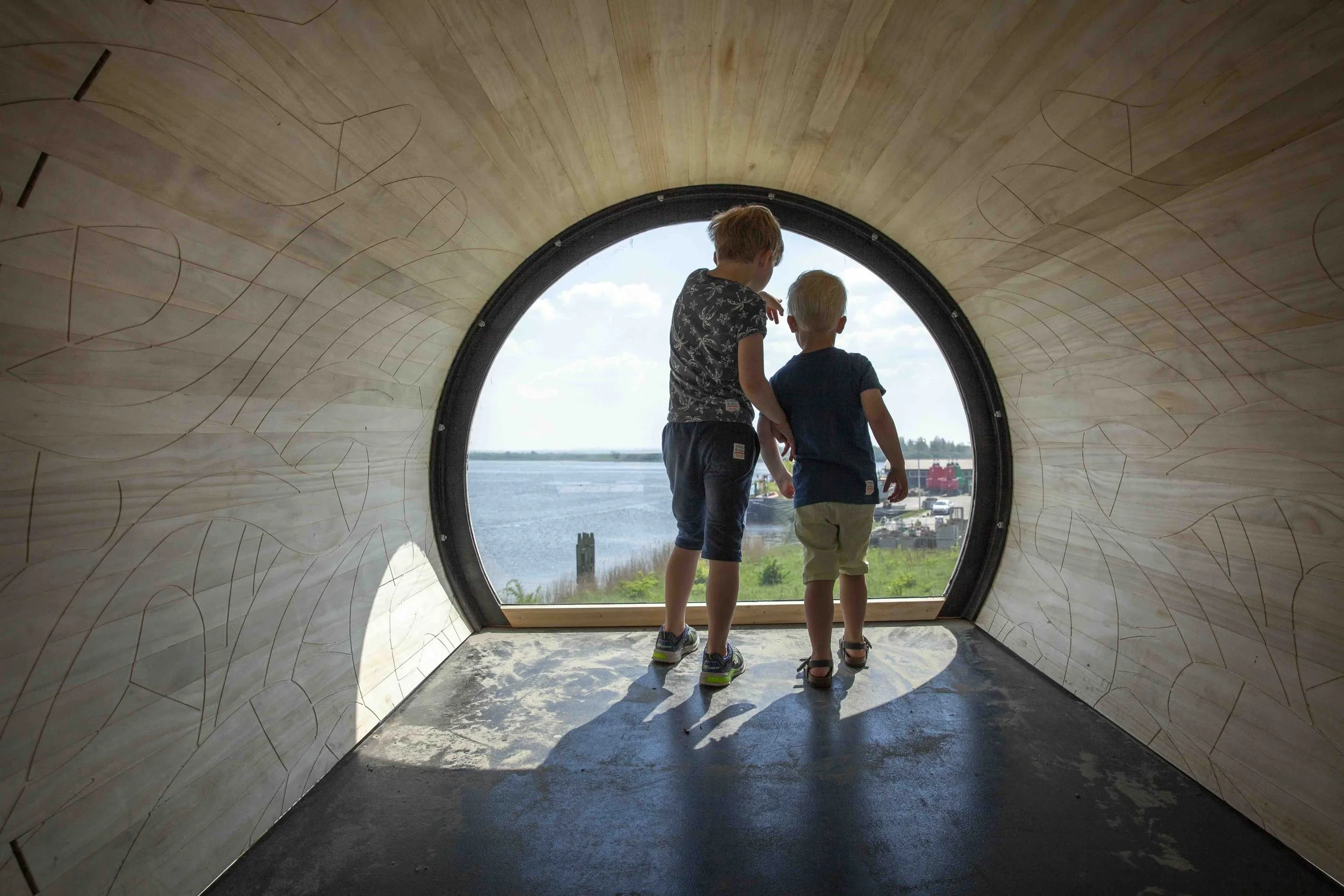 Twee jonge kinderen staan in een ronde tunnel met een houten binnenwand, kijken naar de lucht en het landschap buiten, inclusief water en gebouwen.