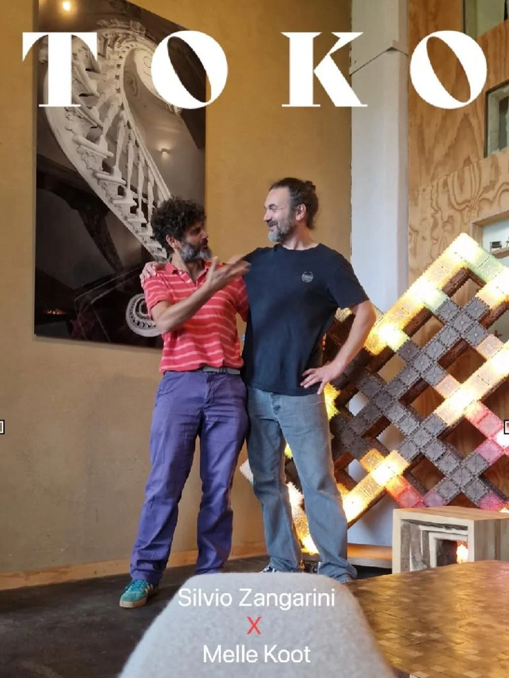 Twee mannen praten en lachen tegen elkaar in een interieur met kunstwerken, een foto en houten decoratie.