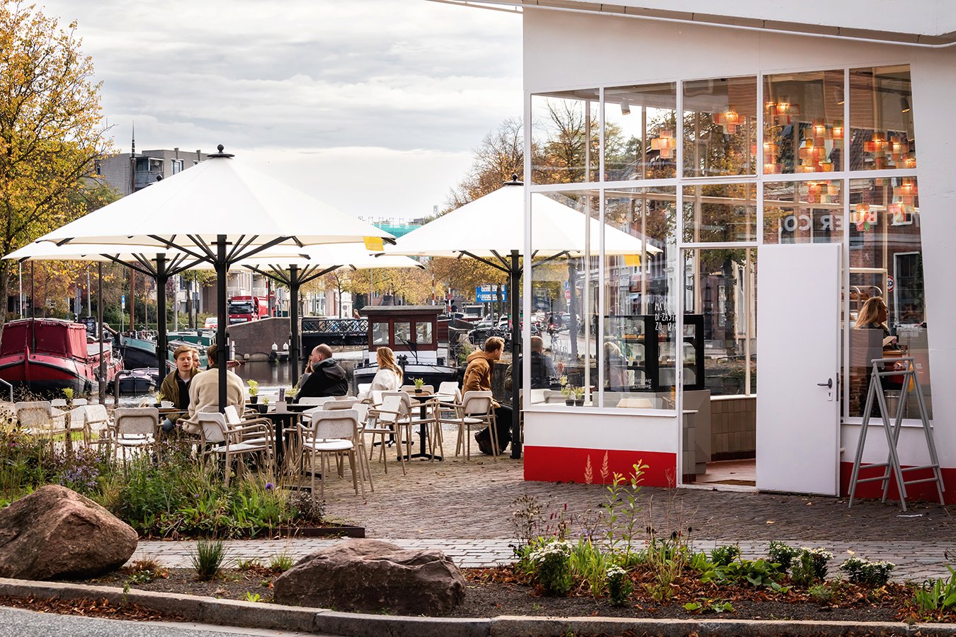 Een terras met witte parasols en witte stoelen buiten een restaurant, gezien langs een kanaal met boten, omgeven door bomen en gebouwen, tijdens de dag.