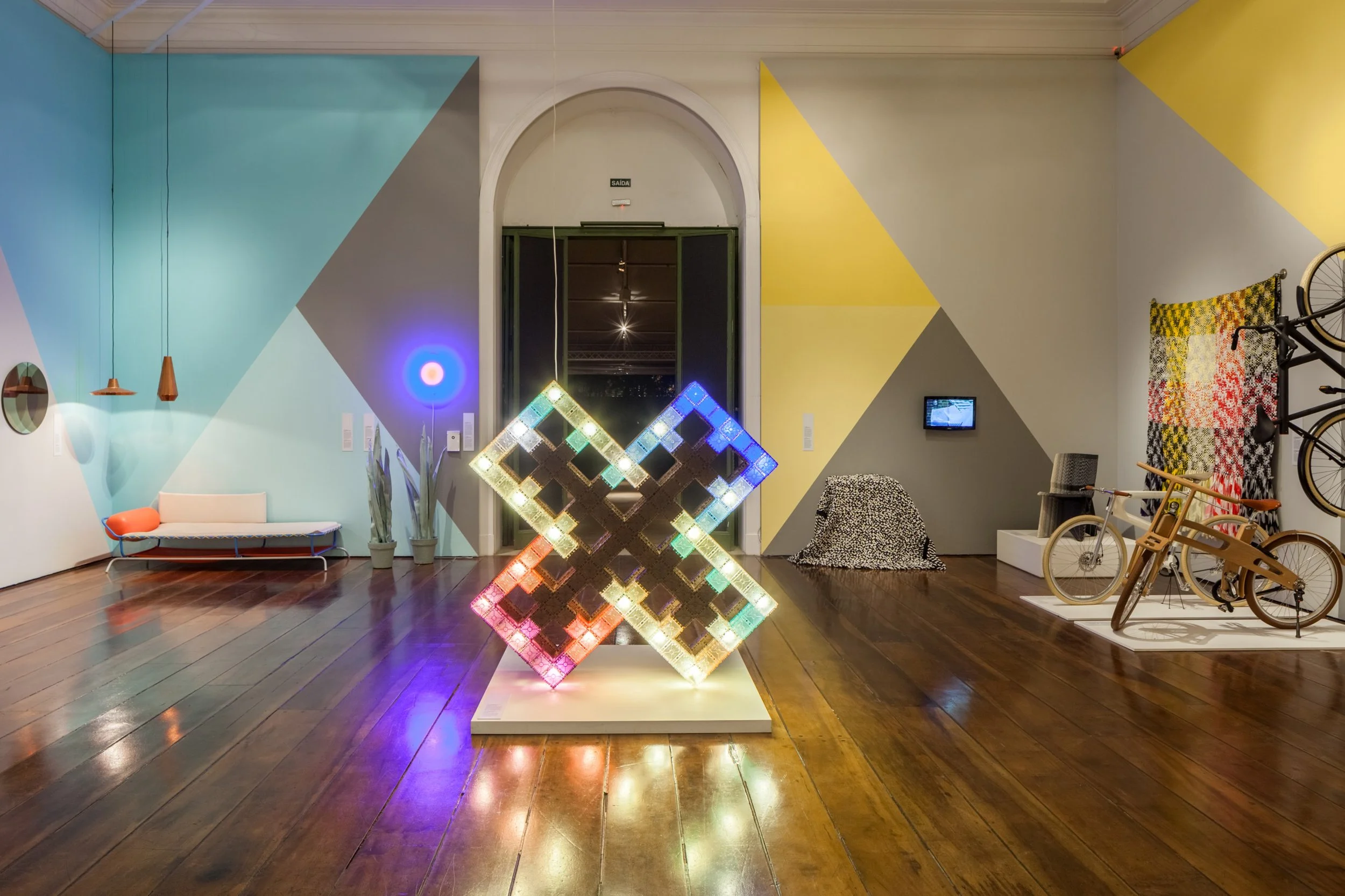 Een kunstzinnige interieurruimtes met kleurrijke geometrische muurkunst, een verlichte digitale installatie, een bank, planten, fietsen, en een muur met een uitgesproken geel en grijs patroon.