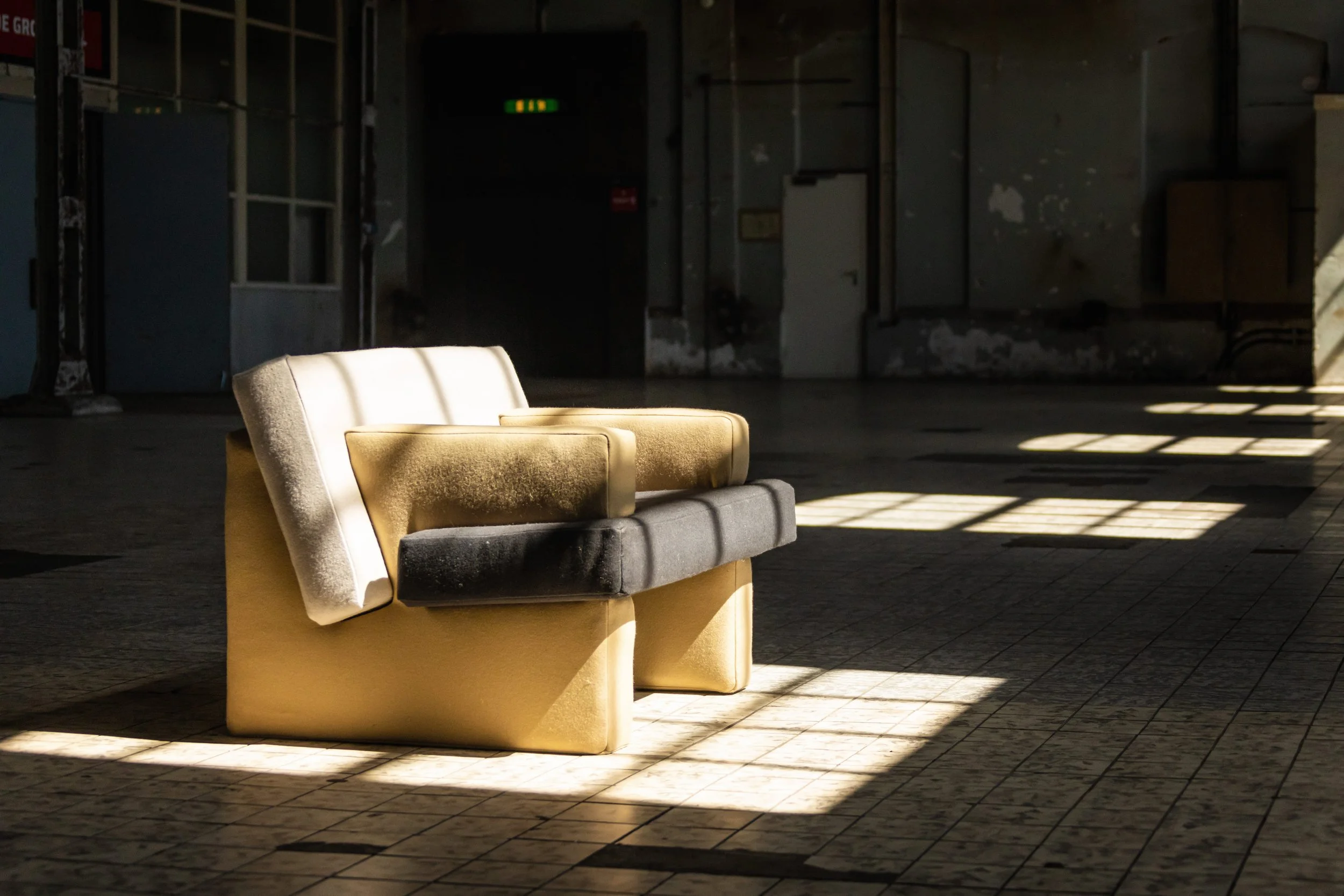 Een beige en grijze bank in een verlaten, verouderde ruimte met gekleurde muren en tegels op de vloer, verlicht door scheve lichtstralen door ramen.