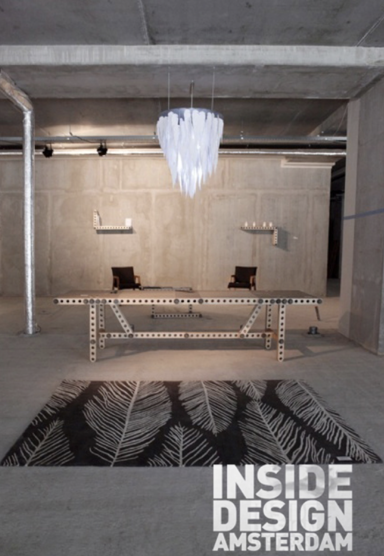 Interieur van een industrieel ogend gebouw met een abstract plafondlamp, twee zwarte stoelen, een tafel met een frame van een bouwpakket, een tapijt met een bladerenpatroon en de tekst 'INSIDE DESIGN AMSTERDAM' op de vloer.