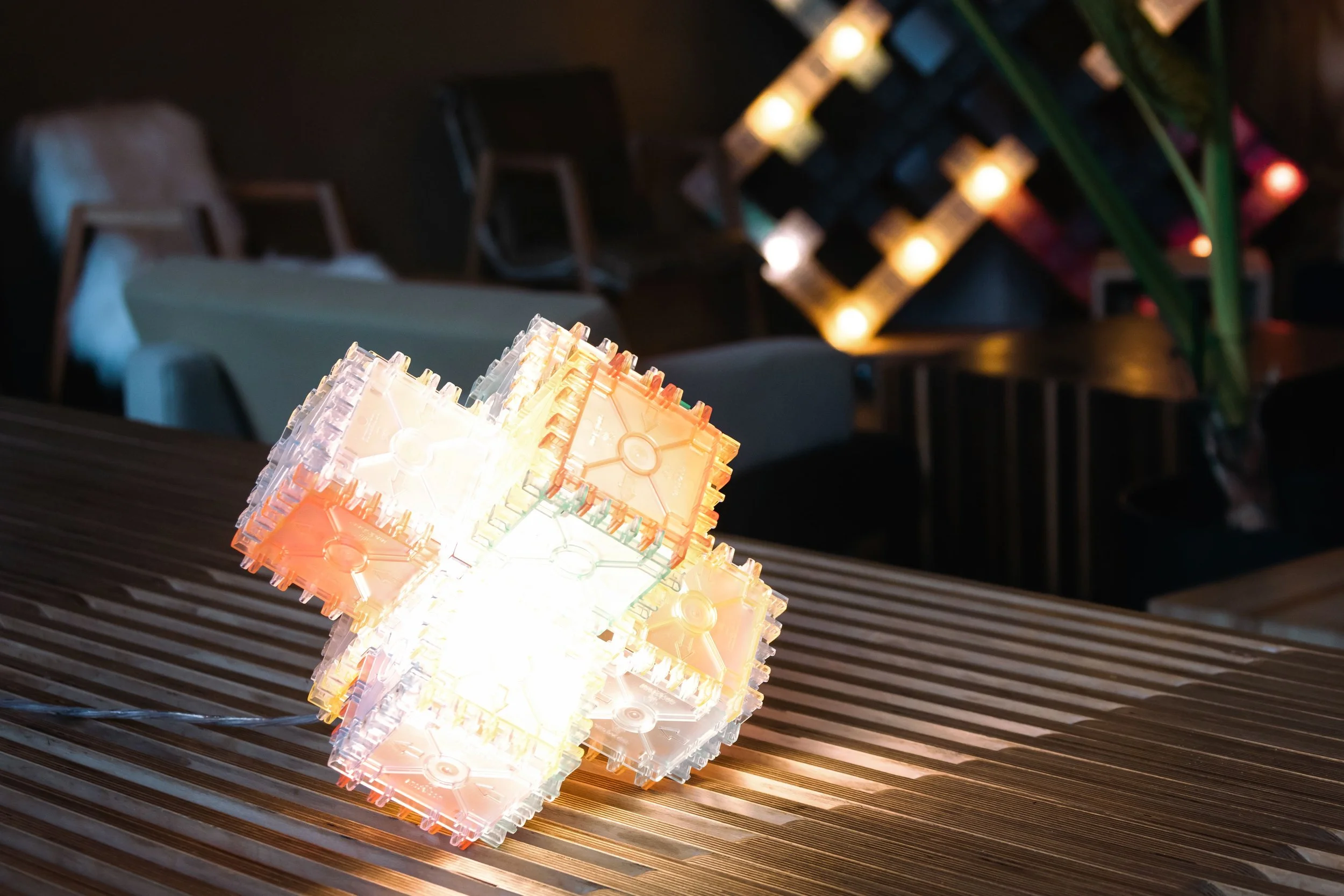 Close-up van een verlichte lamp gemaakt van plastic bouwstenen op een houten tafel, met een achtergrond van andere verlichte bouwelementen en een donkere ruimte.