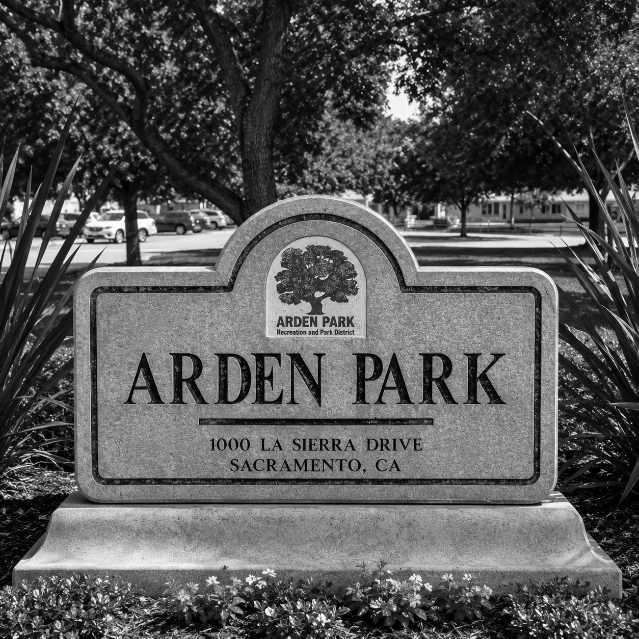 Photograph of a stone sign for Arden Park with the address 1000 La Sierra Drive, Sacramento, CA, surrounded by landscaping and trees, with parked cars and a residential area in the background.