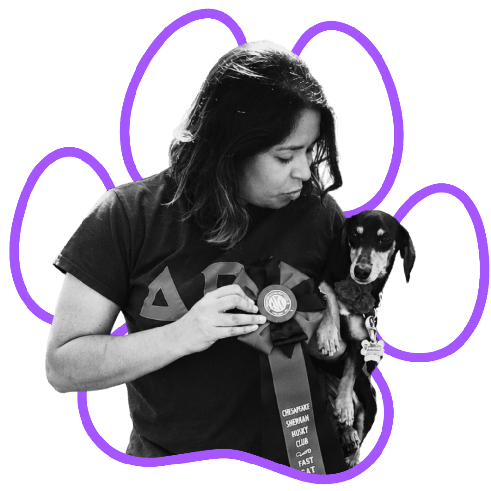 A woman (Jade, Scent Work & Positive Reinforcement Specialist for Wholistic Canine) holding a small dog with a ribbon and medal, against a black background with purple paw print outline around them.