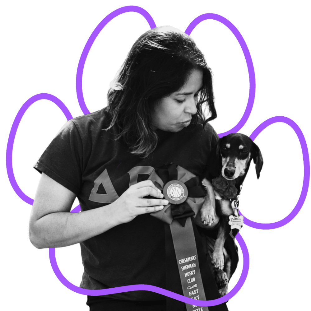 A woman (Jade, Scent Work & Positive Reinforcement Specialist for Wholistic Canine) holding a small dog with a ribbon and medal, against a black background with purple paw print outline around them.