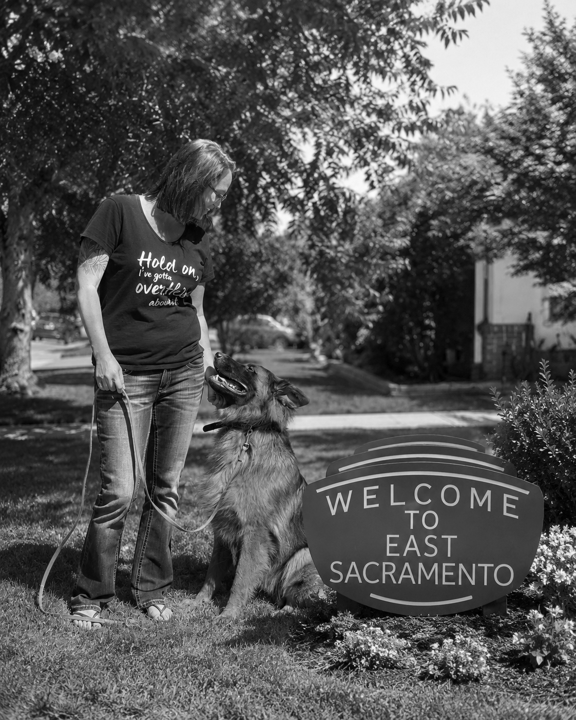 A woman with glasses and a dog sitting next to a welcome sign in East Sacramento on a sunny day.