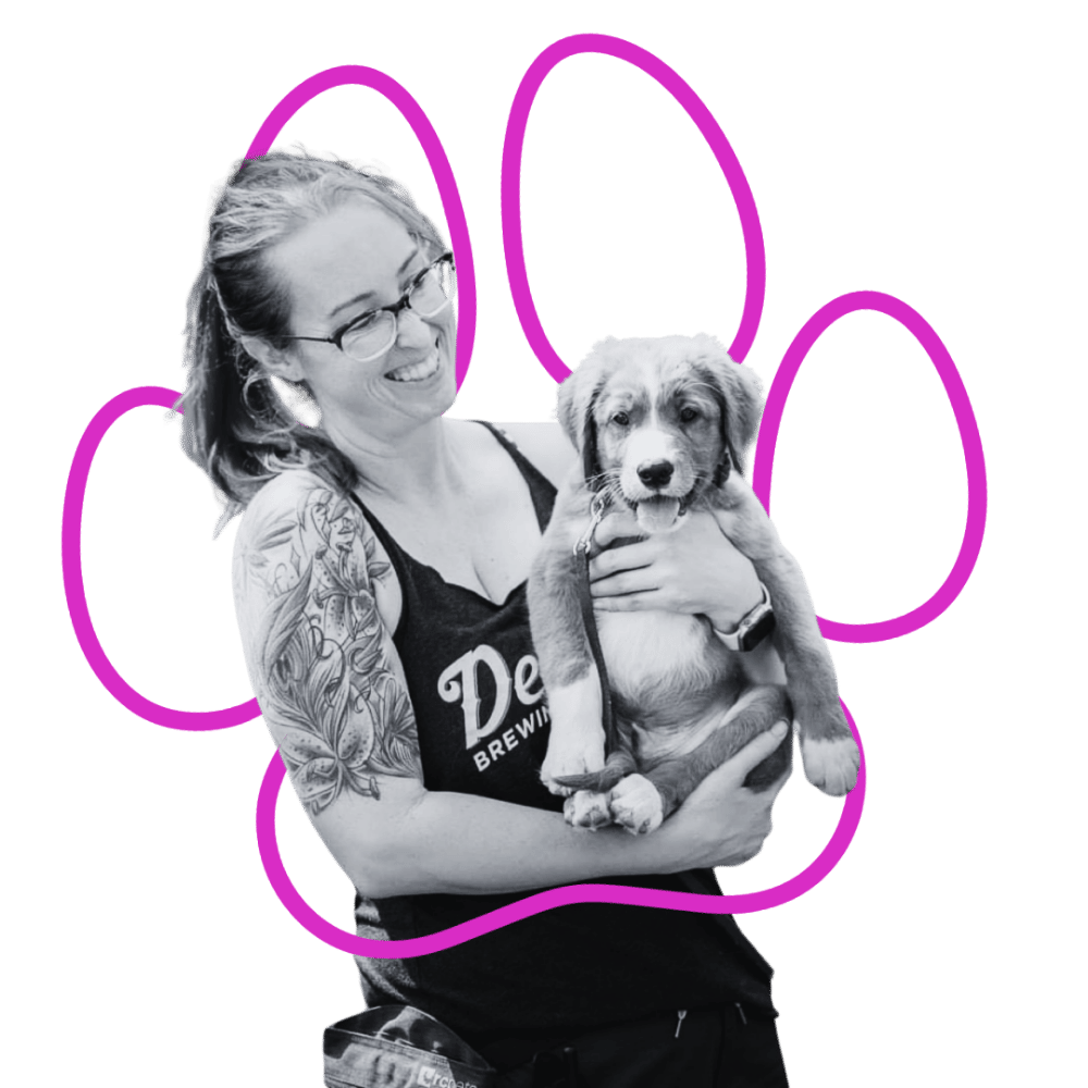 A smiling woman (Terra, Wholistic Canine Owner) with glasses, tattoos, and a black tank top, holding a puppy, with pink paw print outline around her and the puppy like a hug