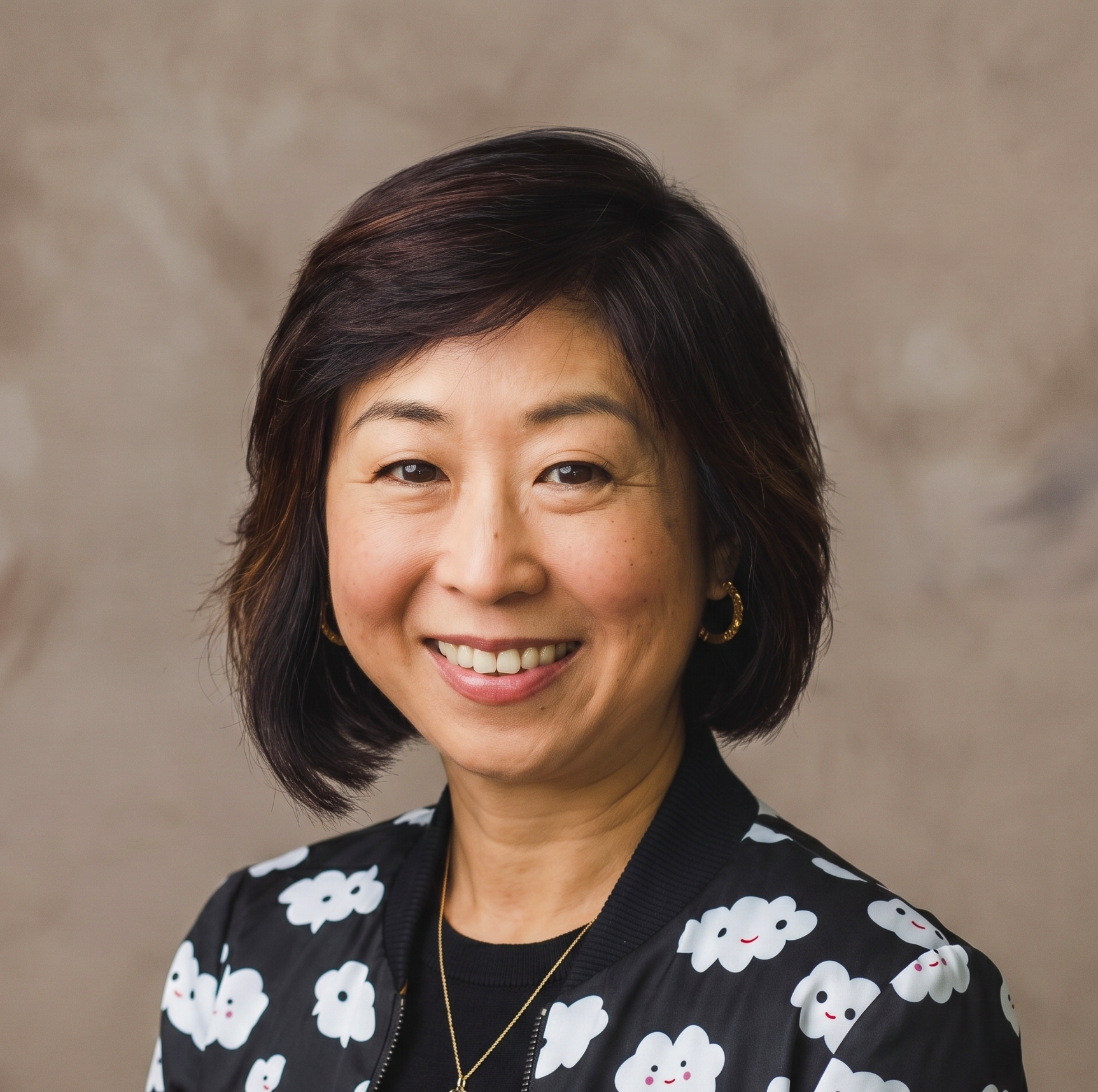 Close-up portrait of a smiling Asian woman with short, dark hair, wearing a black jacket with a white cloud pattern, hoop earrings, and a gold necklace against a neutral background.