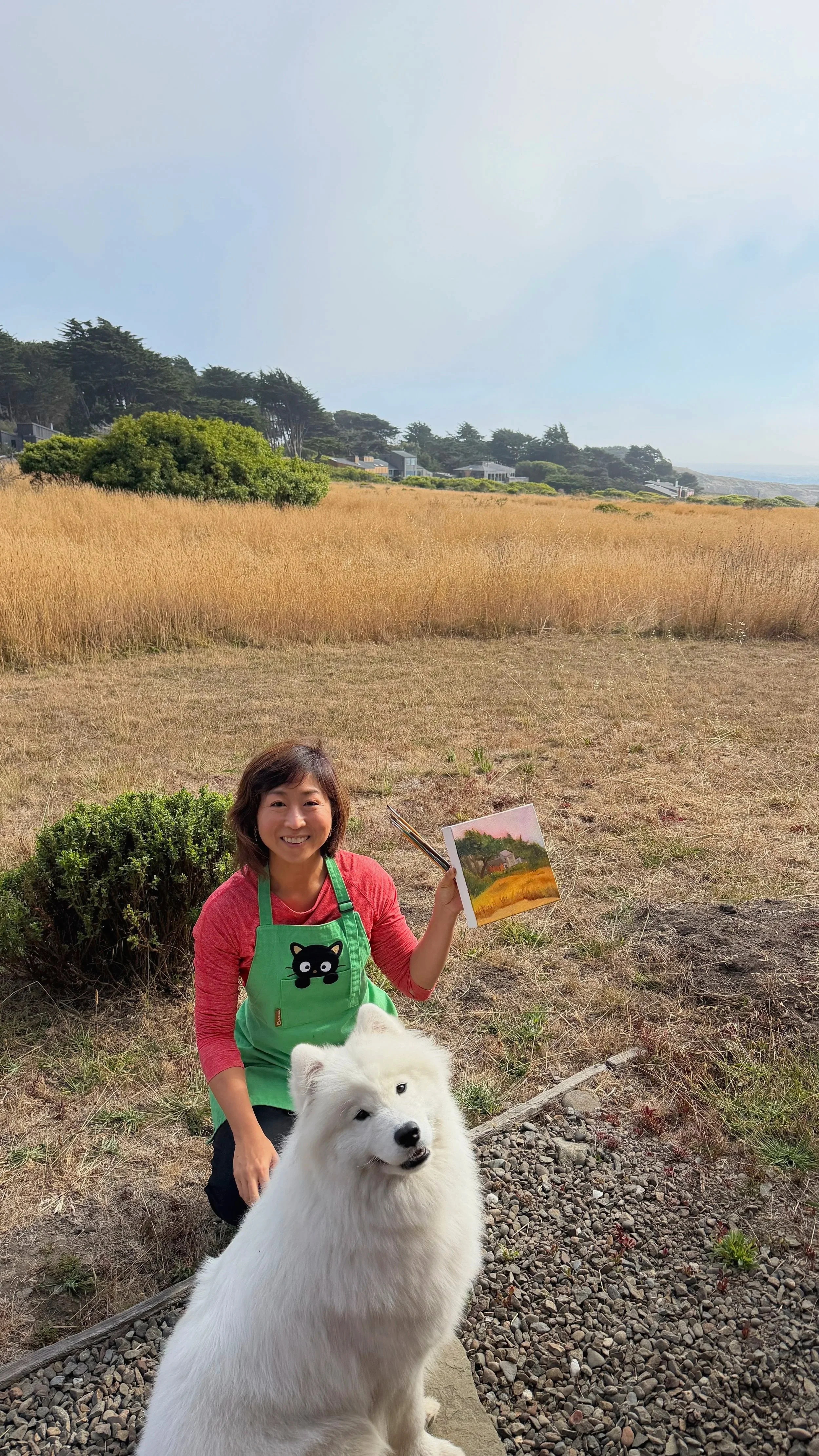 A woman holding a painting of a field, sitting on a grassy area with a large white dog in front of her and a landscape of houses, trees, and a blue sky in the background.