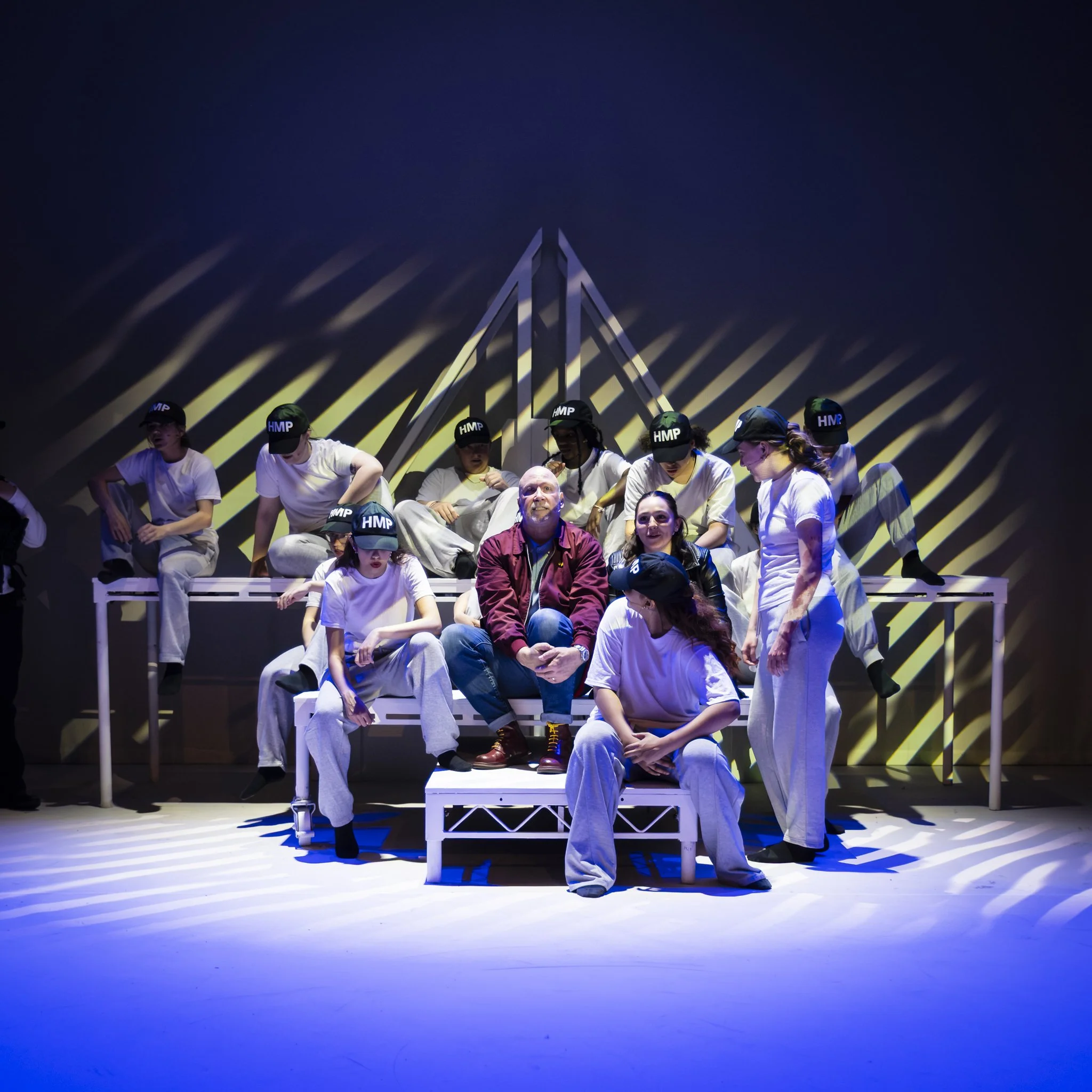 A group of people sitting on a white bench on a stage, with some wearing white and others wearing dark clothing, under dramatic lighting with yellow and blue shadows projected on a dark background.