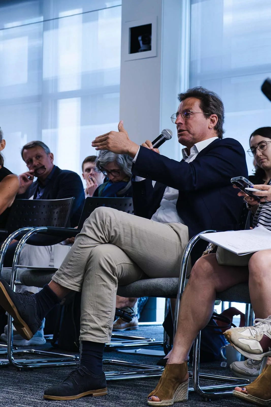 A man in a dark blazer and beige pants is speaking into a microphone at a panel discussion, surrounded by other people.