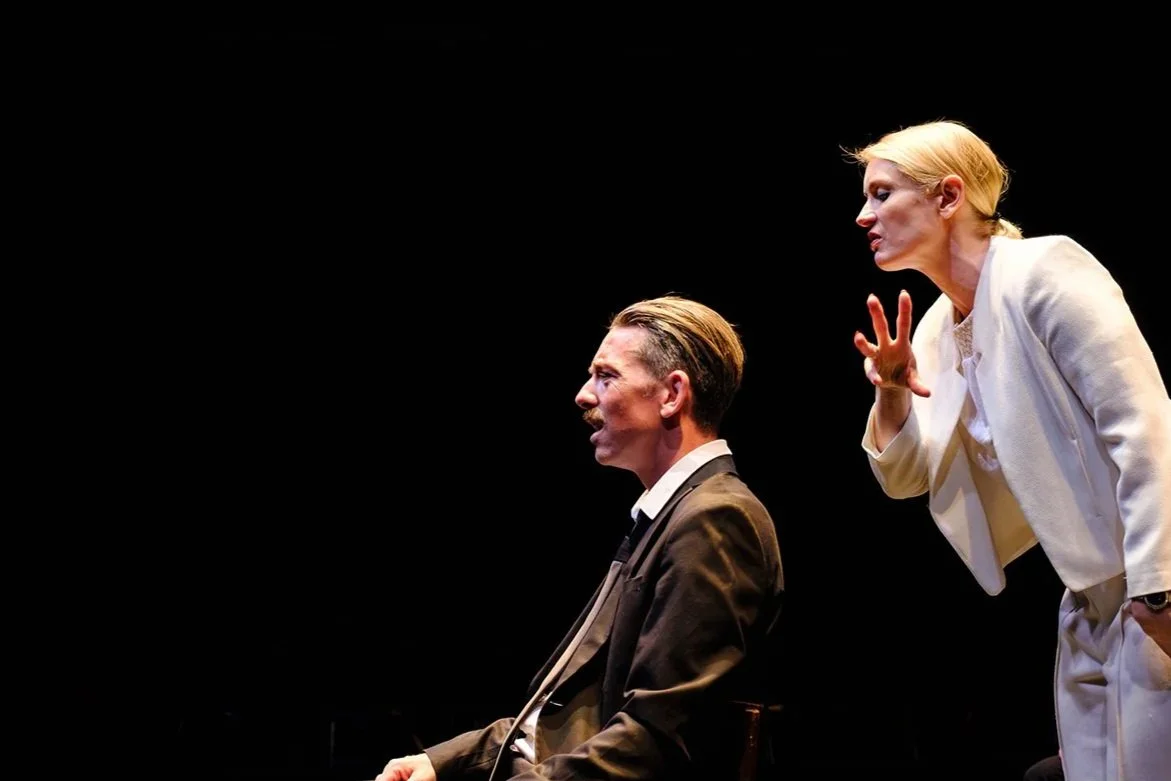 A woman appears to be speaking or arguing with a man who is seated. The woman is wearing white attire and the man is dressed in a brown suit. The background is black.
