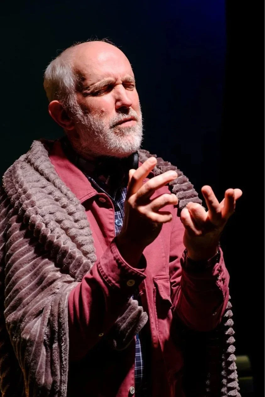 An older man with a beard and closed eyes, appearing to have a contemplative expression, is wearing a reddish-brown jacket with a textured scarf or blanket draped over his shoulders. He is making a gesture with his hands, with fingers slightly apart,