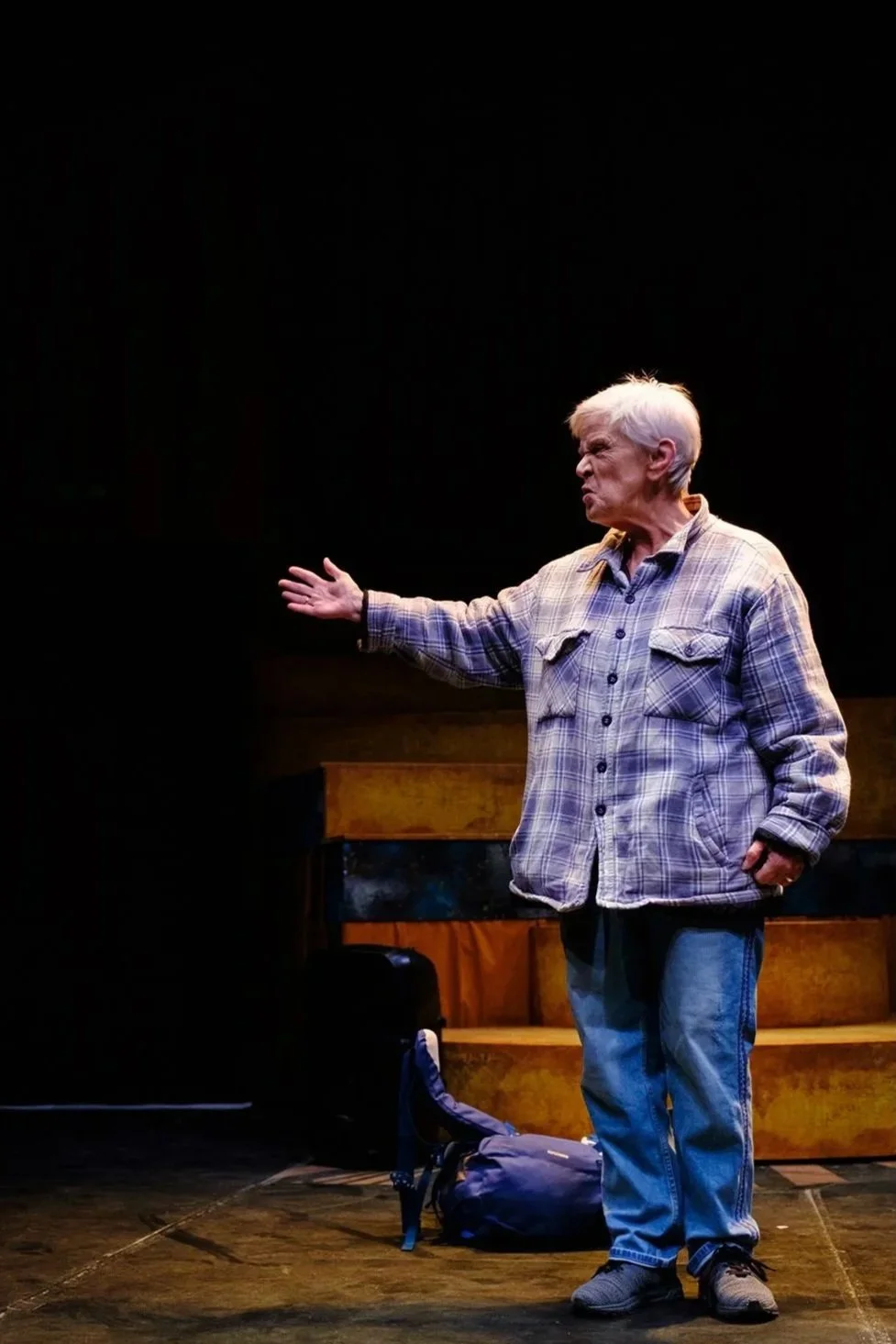 An elderly man with gray hair, wearing a plaid shirt and jeans, appears to be performing on stage with a serious or intense expression, gesturing with his right arm extended.