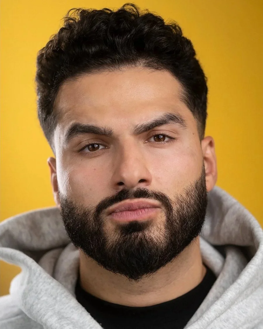 Close-up portrait of a young man with dark, curly hair, a full beard, wearing a gray hoodie, against a bright yellow background.