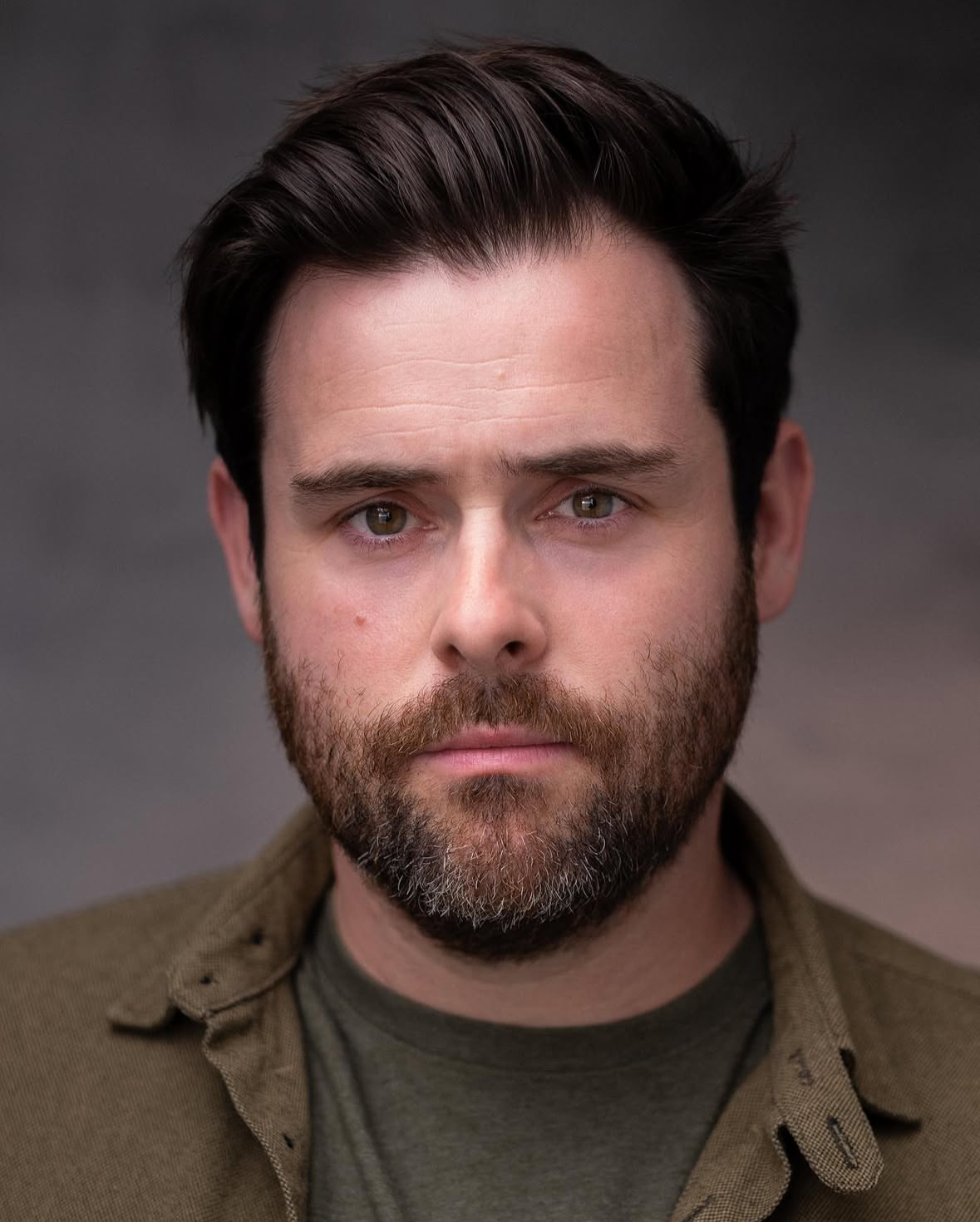 Close-up portrait of a man with brown hair and a beard, wearing a brown jacket and a dark shirt, looking directly at the camera.