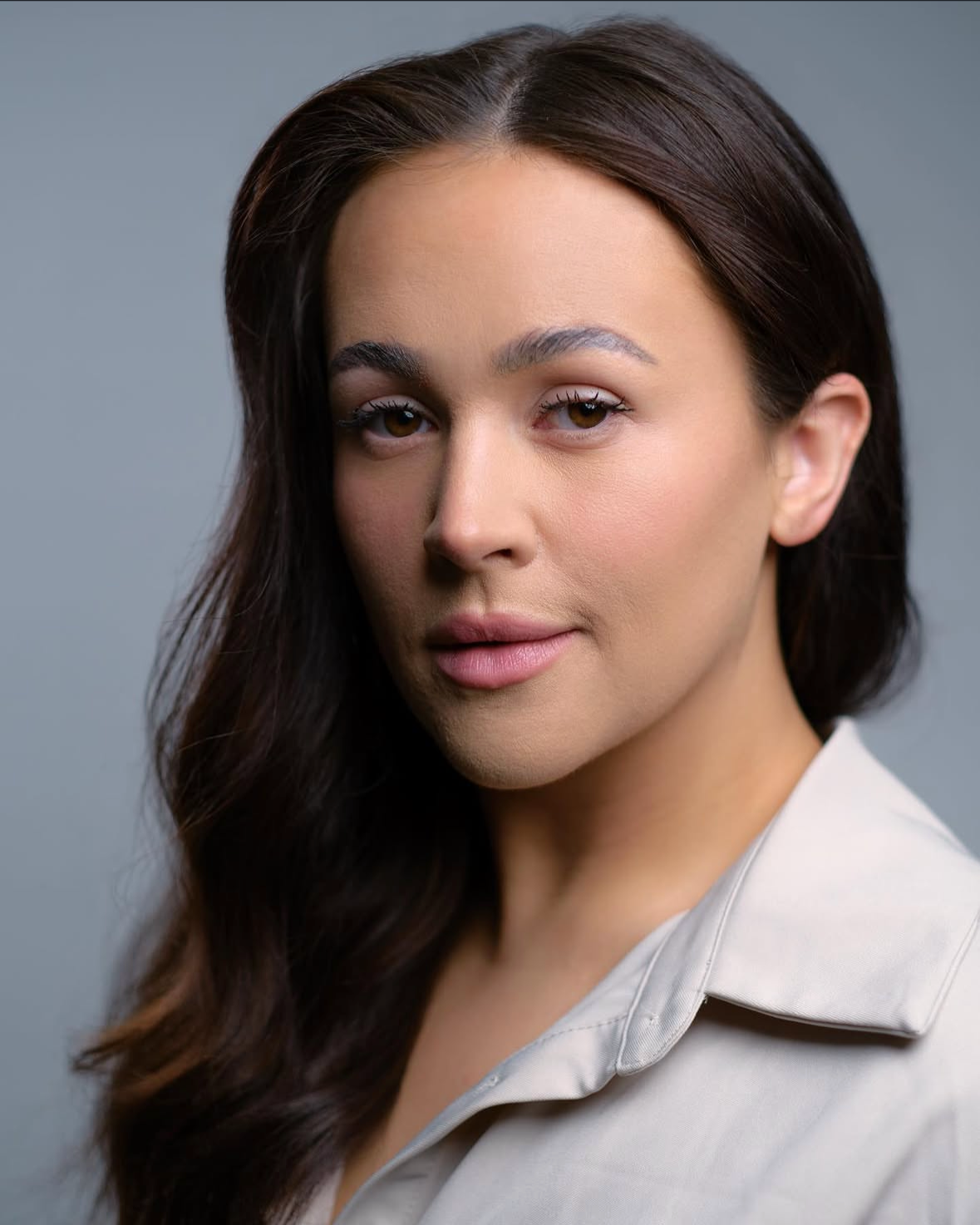 Close-up portrait of a woman with dark brown hair and light makeup, wearing a light-colored shirt, against a gray background.